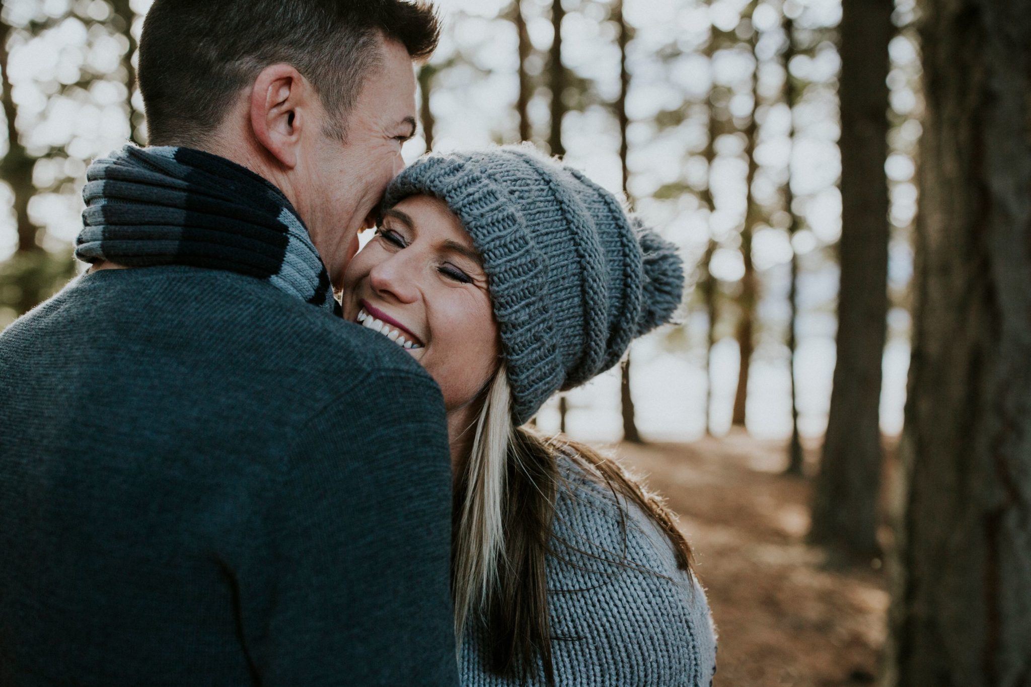 Charlotte Kiri Photography - Engagement Photography with a happy couple smiling wearing winter knitwear, and embracing in the trees of a forest in Wanaka, New Zealand