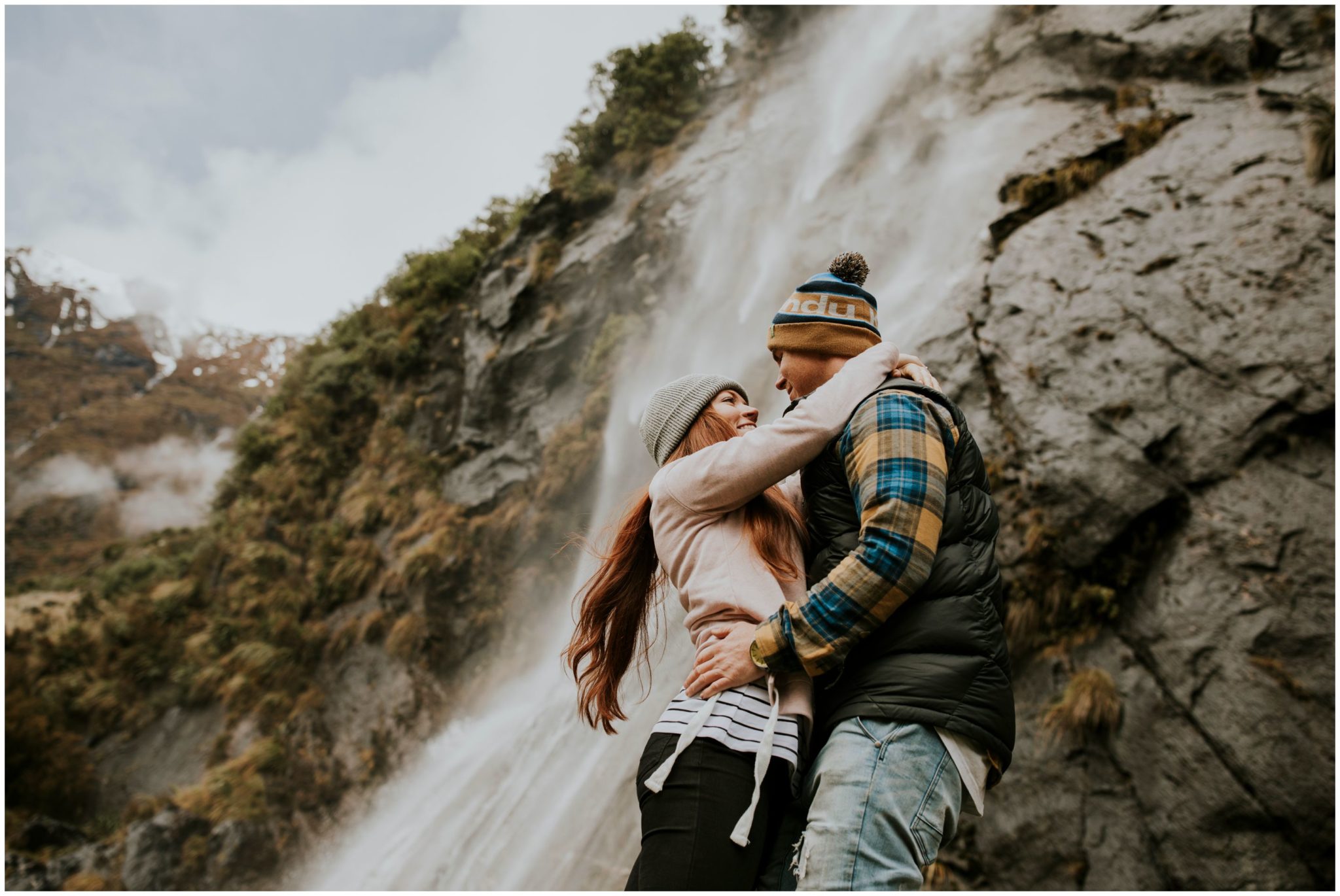 Charlotte Kiri Photography - Engagement Photography with a gorgeous couple embracing and smiling at each other, next to a misty romantic waterfall, in Wanaka, New Zealand