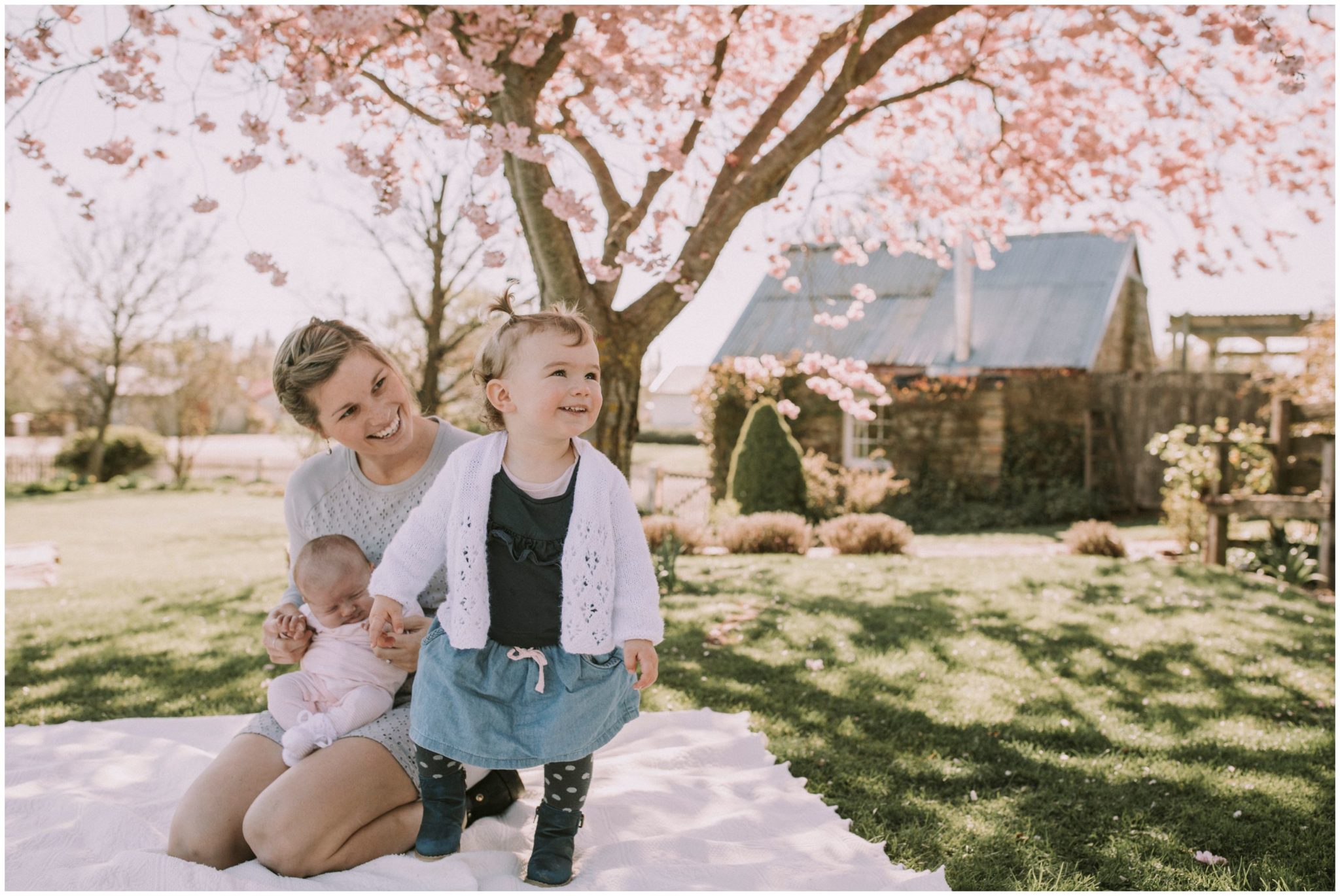 Charlotte Kiri Photography - Family Photography of a newborn baby girl being held on her mothers lap on a picnic blanket, while her toddler sister stands in front of them smiling, with a pink blossom tree and pretty cottage behind in Wanaka, New Zealand.