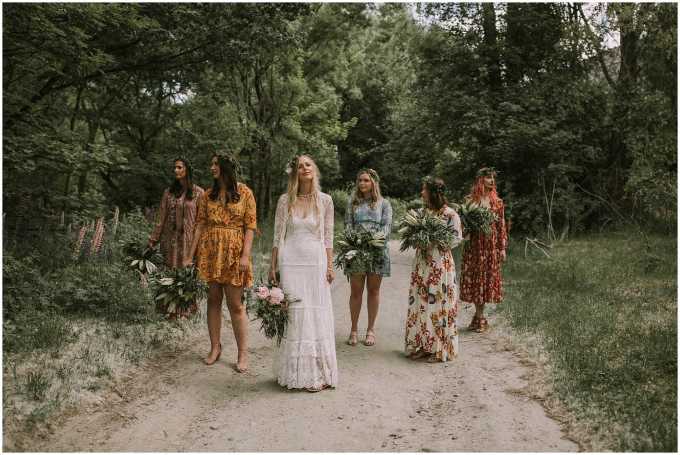 Charlotte Kiri Photography - Wedding Photography of a bride wearing a pretty bohemian lace dress standing on a secluded dirt road and surrounded by her lovely bridesmaids who wear colourful floral dresses with wreaths and rustic bouquets, in Arrowtown, New Zealand.