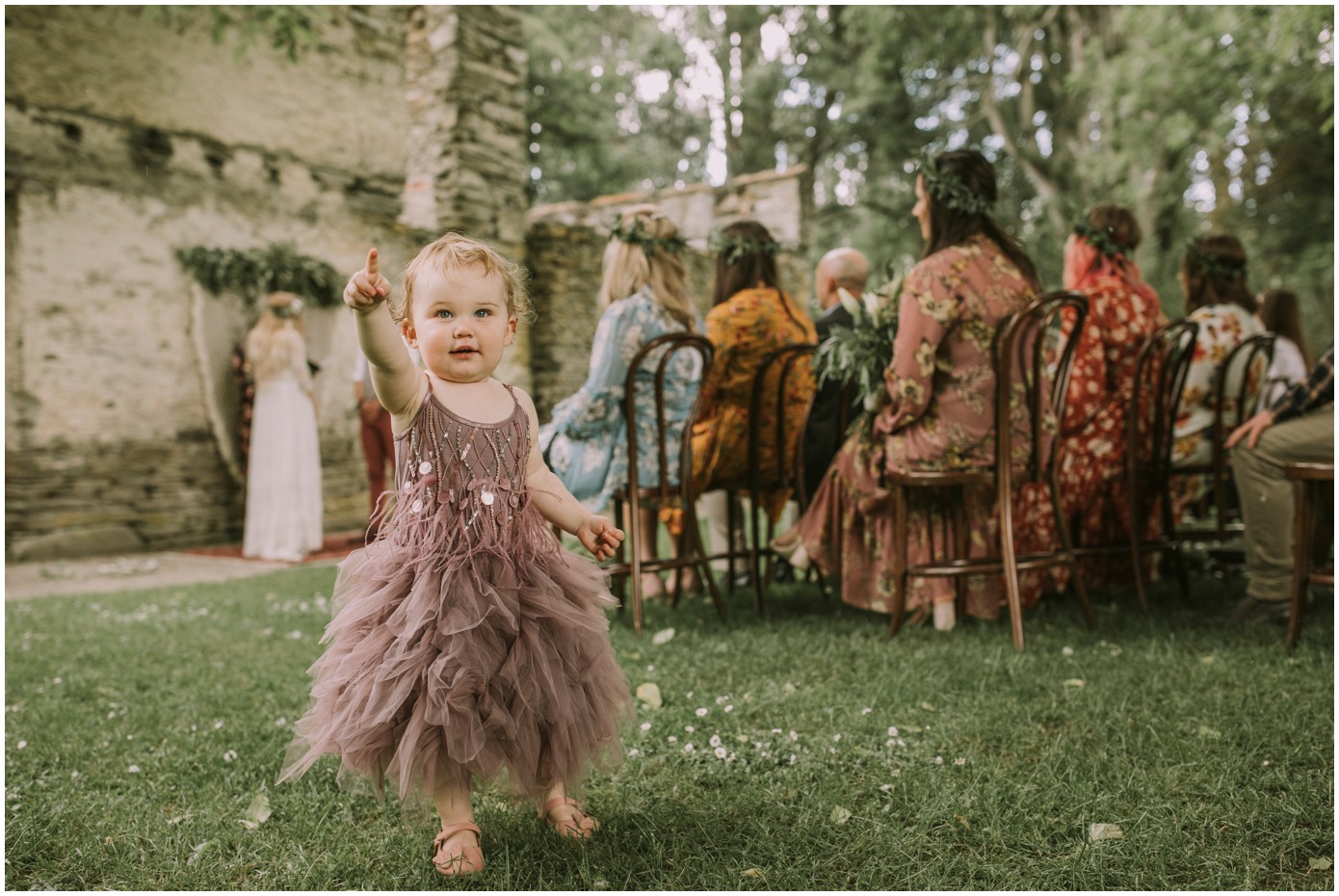 Charlotte Kiri Photography - Wedding Photography of a toddler wearing a muted plum dress and pointing at the camera, as the bride and groom exchange vows at the alter at Thurlby Domain in Arrowtown, New Zealand.