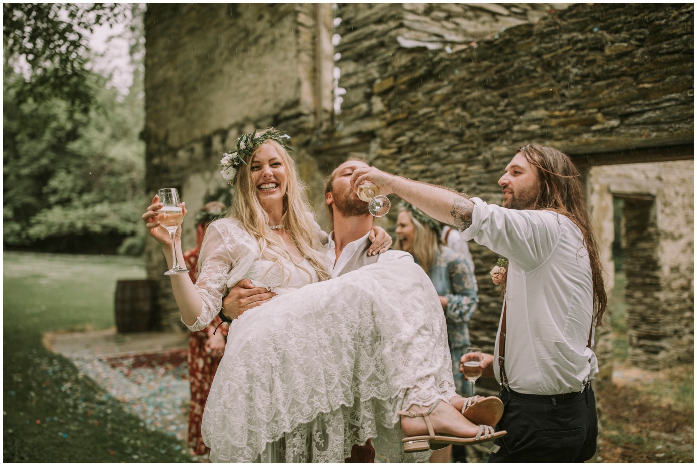 Charlotte Kiri Photography - Wedding Photography of a bohemian bride wearing a pretty lace dress, who is laughing, as her groom lifts her up, as they celebrate and drink champagne outside a rustic building at Thurlby Domain, Arrowtown, New Zealand.