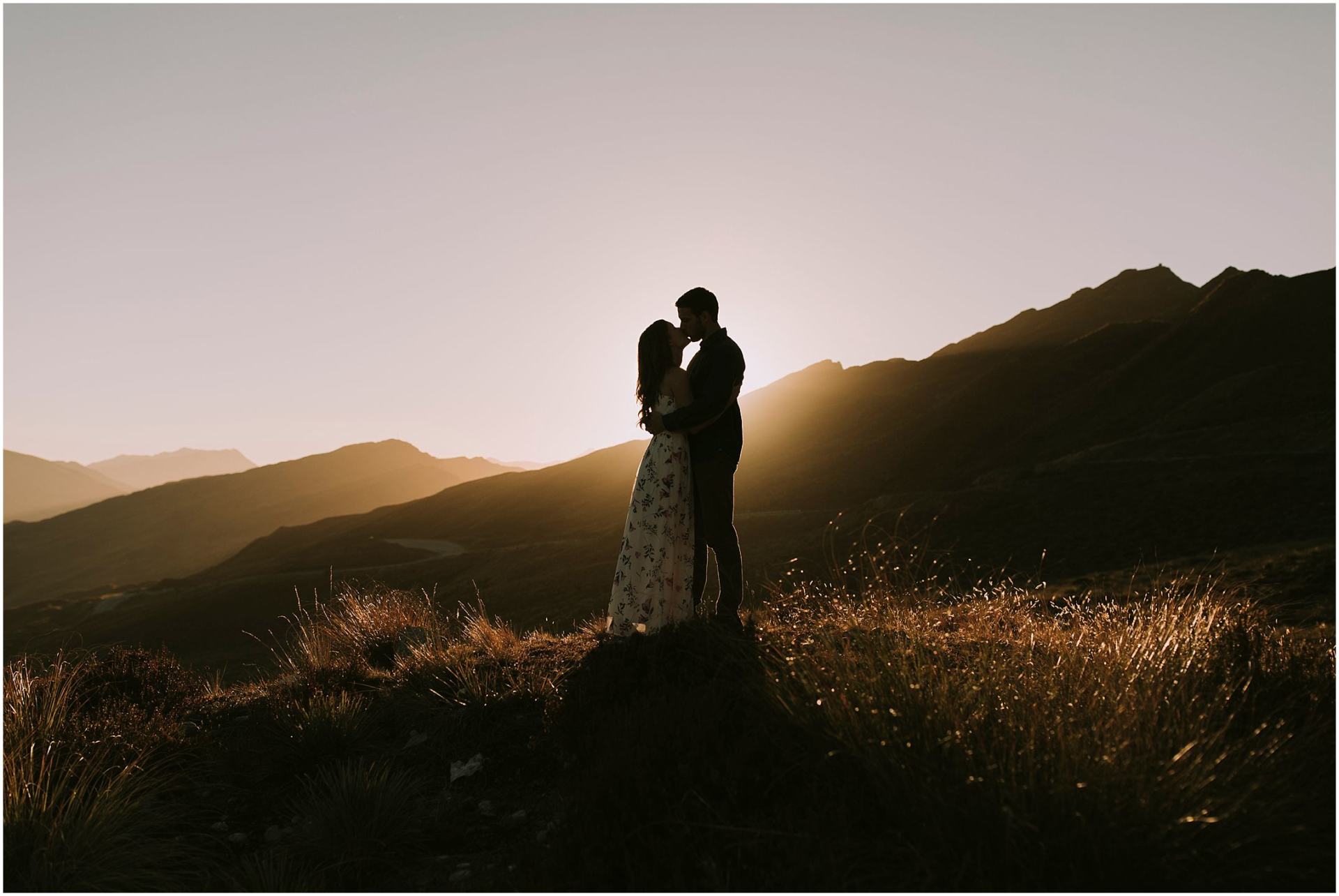 Charlotte Kiri Photography - Engagement Photography with an admiring couple standing on a clifftop in silhouette, with the sun setting behind the mountains in the distance in Wanaka, New Zealand