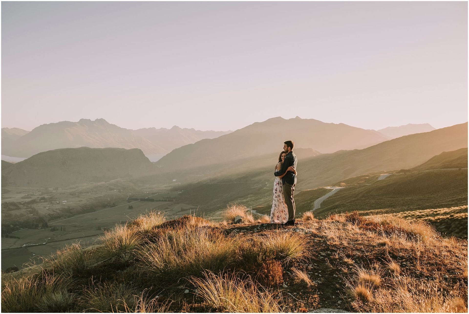 Charlotte Kiri Photography - Engagement Photography with a couple embracing in the sunset, standing on top of a hill overlooking beautiful landscape with mountains and lakes behind in Wanaka, New Zealand
