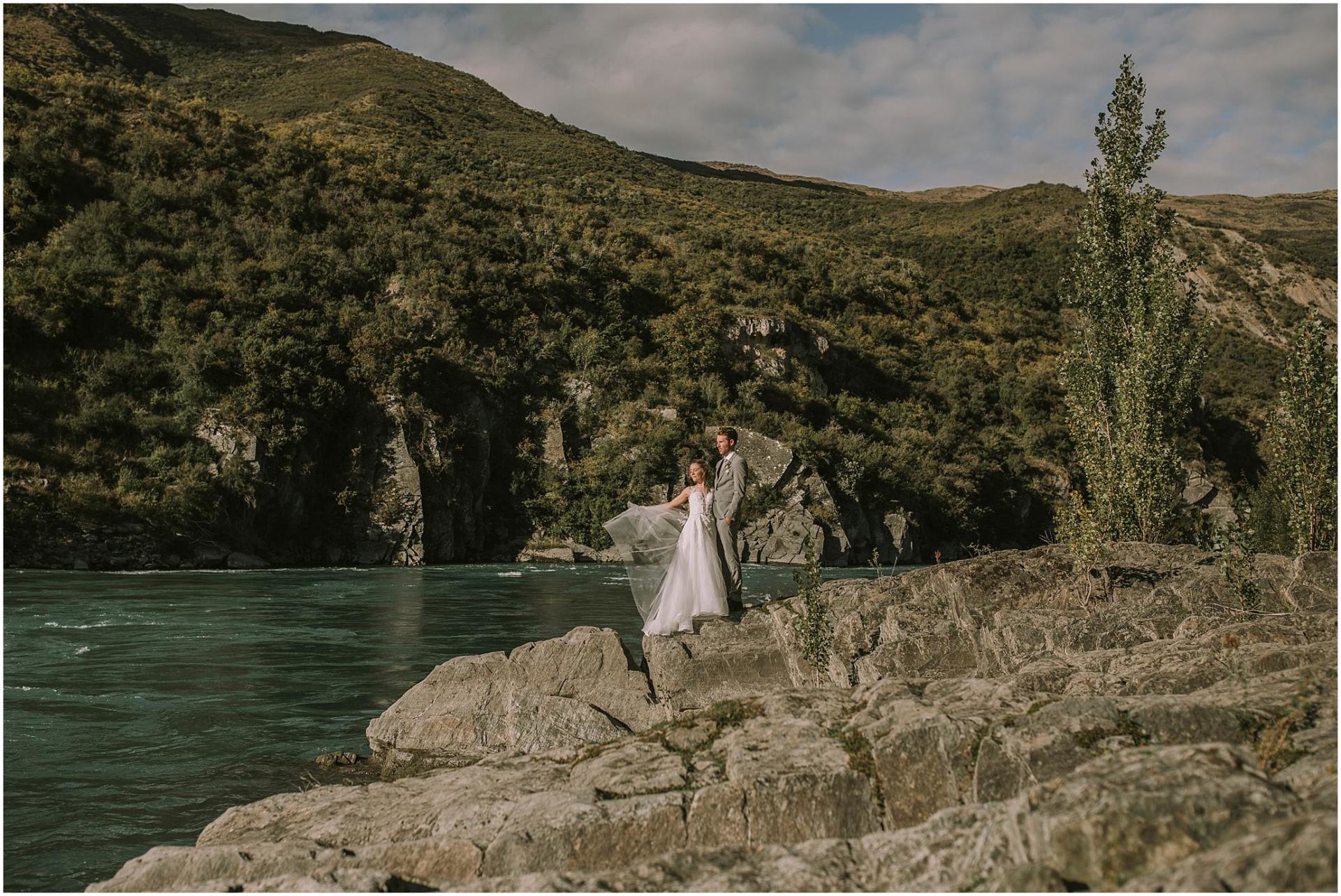 Charlotte Kiri Photography - Wedding Photography of a bride wearing a pretty strapless gown and throwing her veil into the wind, as she stands next to her groom who is wearing a smart grey suit, as they stand on rocks overlooking a stunning lake with mountains behind in Queenstown, New Zealand.