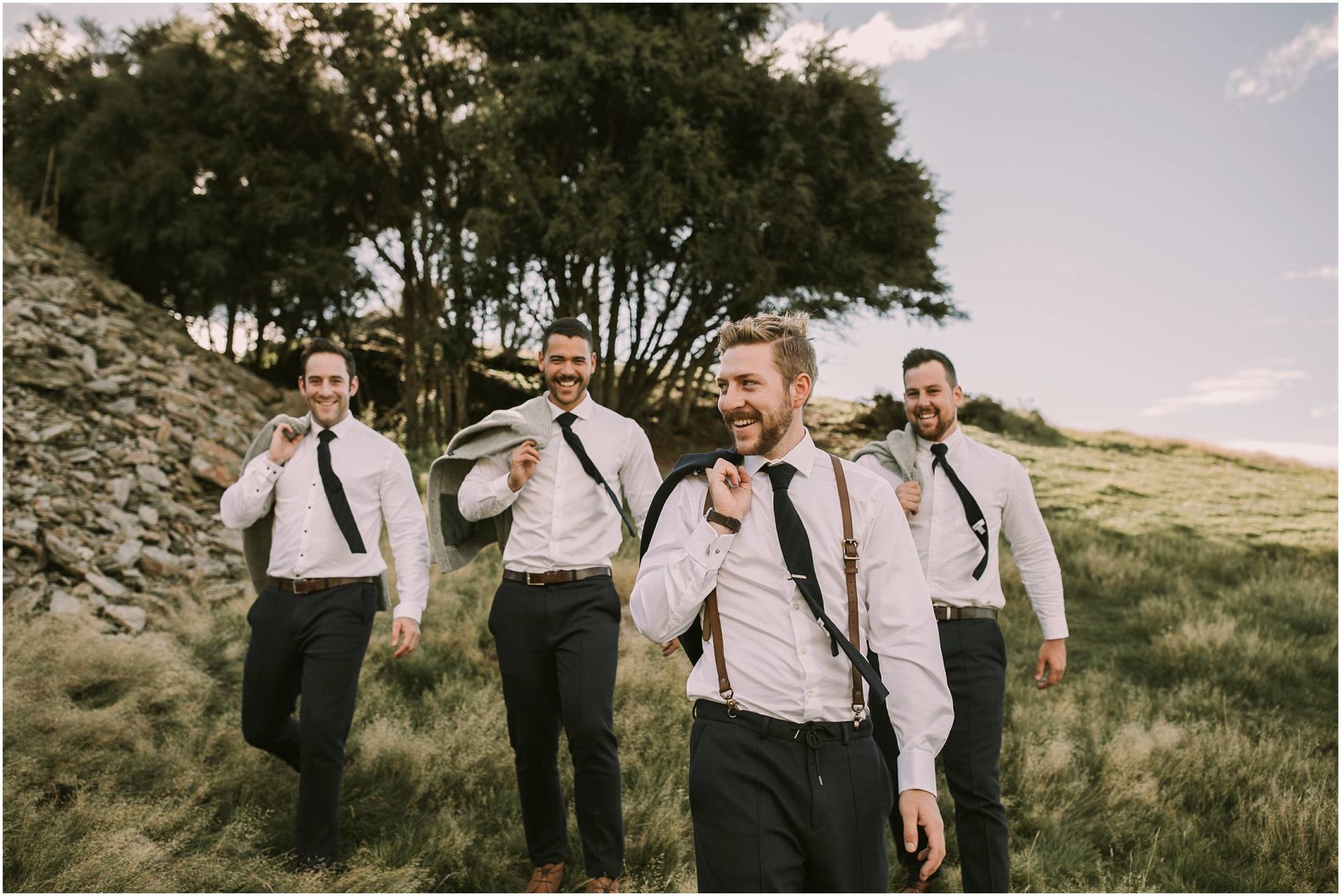 Charlotte Kiri Photography - Wedding Photography of a smart groom and groomsmen, smiling and wearing black pants with black tie and braces, and their jackets over their shoulders, whilst walking in the long grass near Criffel Station, Wanaka, New Zealand.
