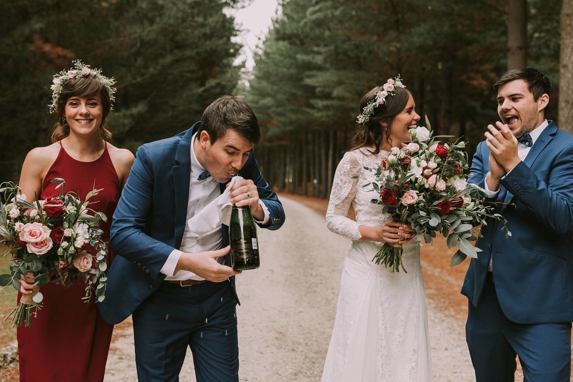 Charlotte Kiri Photography - Wedding Photography of a bride wearing a floral wreath and long-sleeved vintage dress and groom wearing a steel blue suit, and members of the bridal party smiling and drinking champagne as they walk down a gravel road lined with trees at Criffel Station, Wanaka, New Zealand.