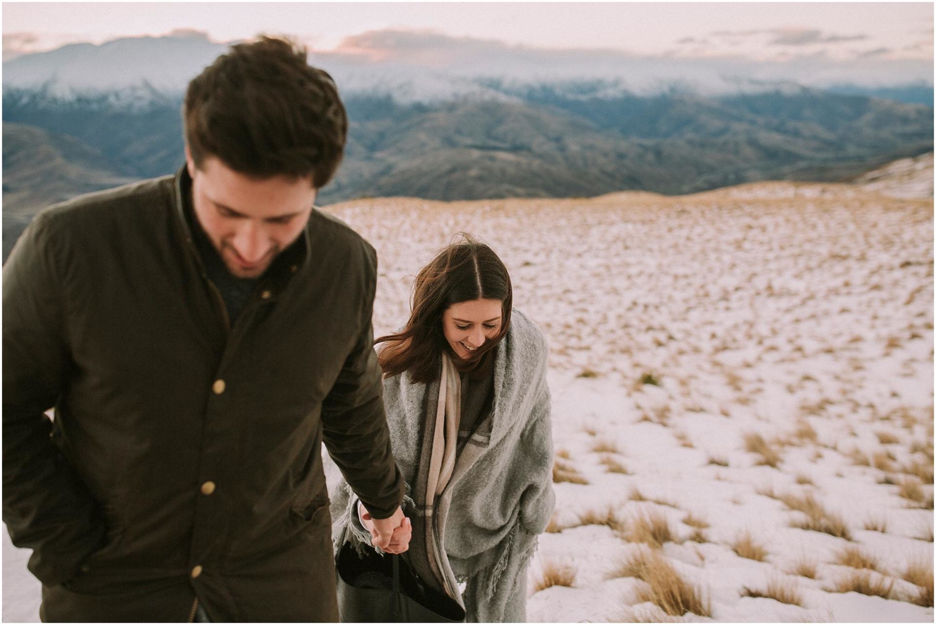 Charlotte Kiri Photography - Engagement Photography with a happy couple holding hands and smiling as they walk over snow-covered tussock grass, with mountains behind, in Wanaka, New Zealand