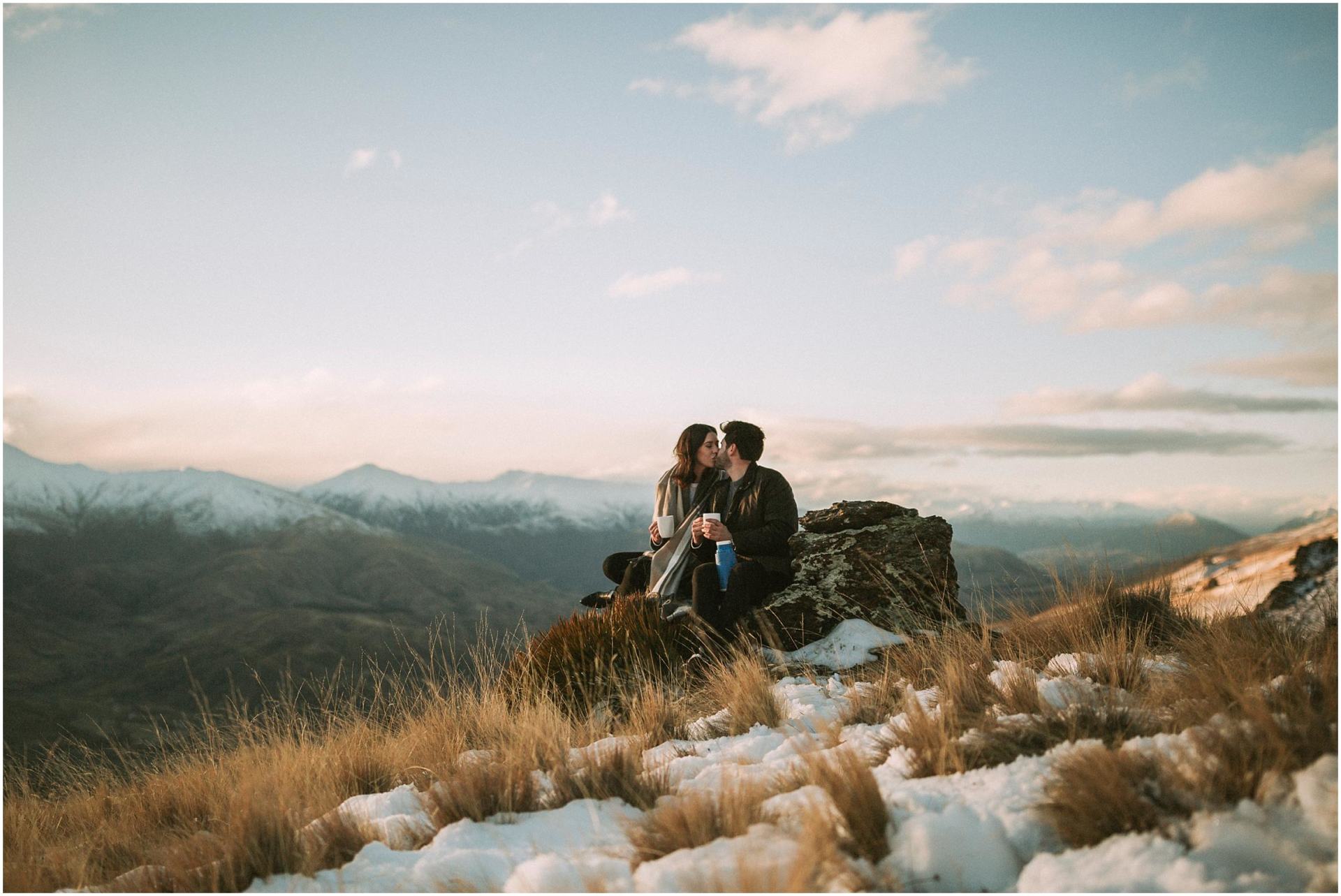 Charlotte Kiri Photography - engagement photography with a cosy couple kissing and holding their coffee, whilst sitting on a rock on the edge of a rugged mountain, amoungst the snow and tussock grass, with a backdrop of stunning snow-capped mountains behind, in Wanaka, New Zealand