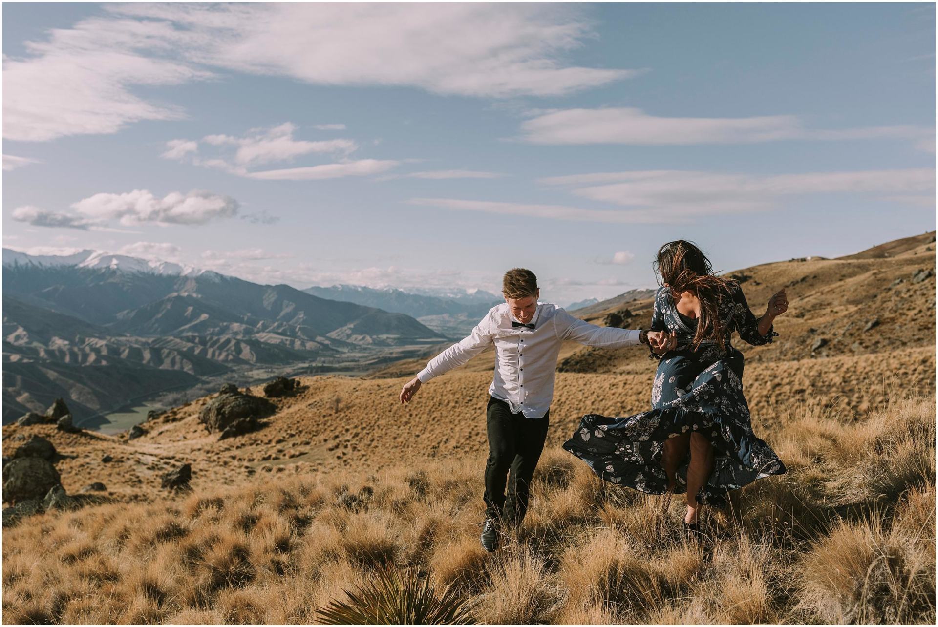 Charlotte Kiri Photography - Engagement Photography with a joyous couple holding hands and running in the tussock grass on a mountain with the stunning landscape of mountains and valleys behind in Wanaka, New Zealand
