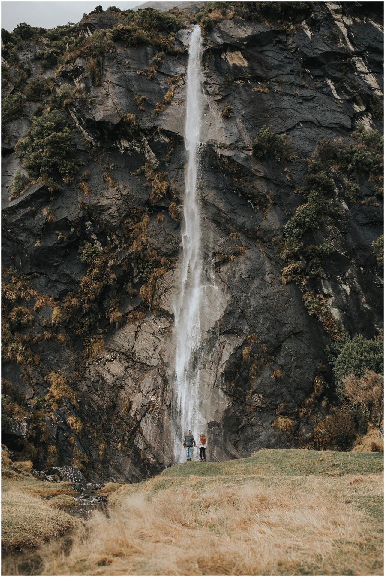Charlotte Kiri Photography - Engagement Photography with a stunning image of a couple standing looking up at a waterfall cascading over a jagged rockface, in Wanaka, New Zealand
