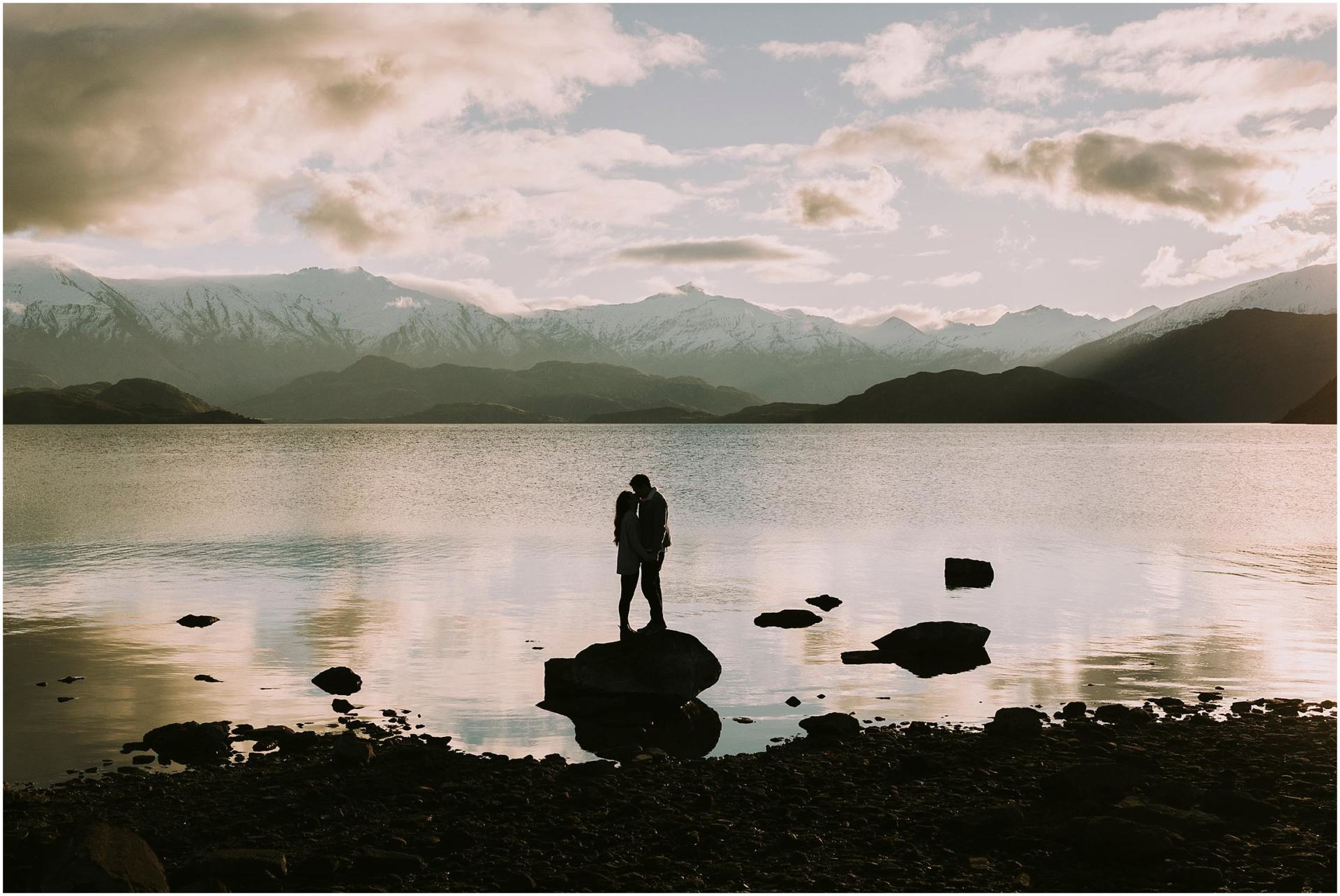 Charlotte Kiri Photography - Engagement Photography with a romantic silhouette shot of a couple standing kissing on a rock in front of a picturesque lake with snow-capped mountains behind in Wanaka, New Zealand