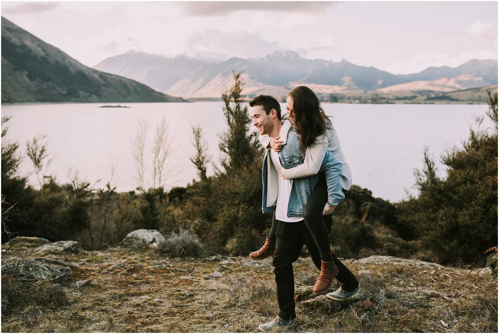 Charlotte Kiri Photography - Engagement Photography with a happy woman taking a piggyback ride on her smiling partner in front of a stunning lake with gorgeous mountainous terrain behind in Wanaka, New Zealand
