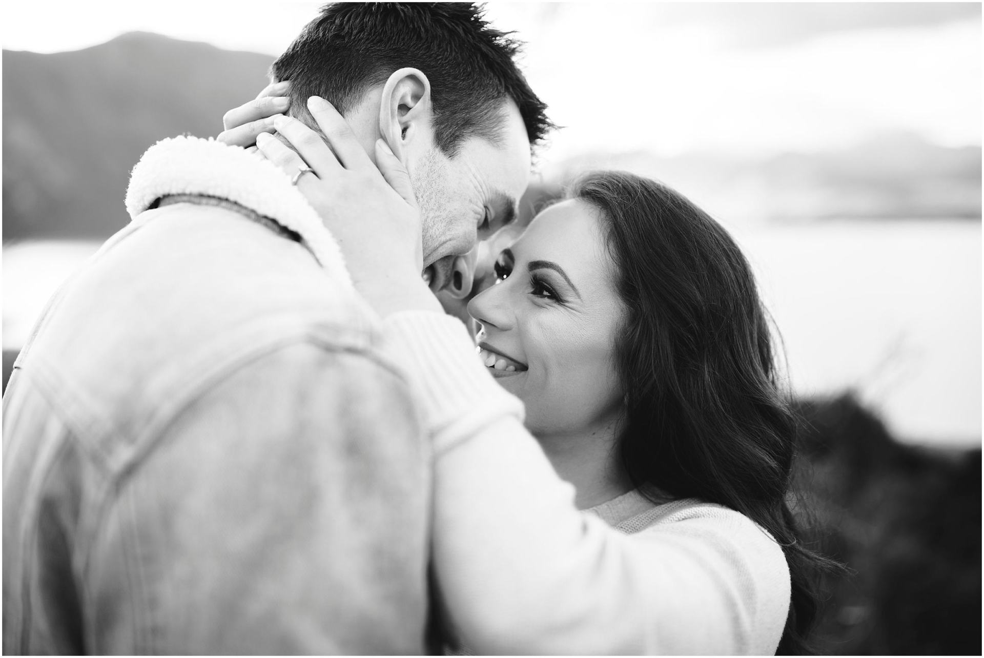Charlotte Kiri Photography - Engagement Photography with a black and white close-up of a happy couple embracing and smiling at each other, with a lake and mountains faded out in the background, in Wanaka, New Zealand