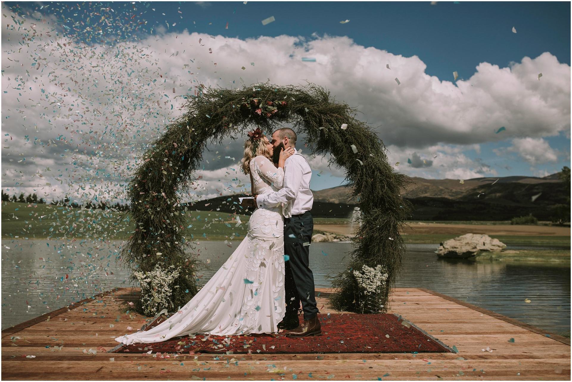 Charlotte Kiri Photography - Wedding Photography of a bride and groom standing at the end of a jetty as confetti is scattered around them, as they kiss and embrace lovingly, under an arch made of rustic green foliage and white flowers, with a backdrop of mountains and fields in Wanaka, New Zealand.