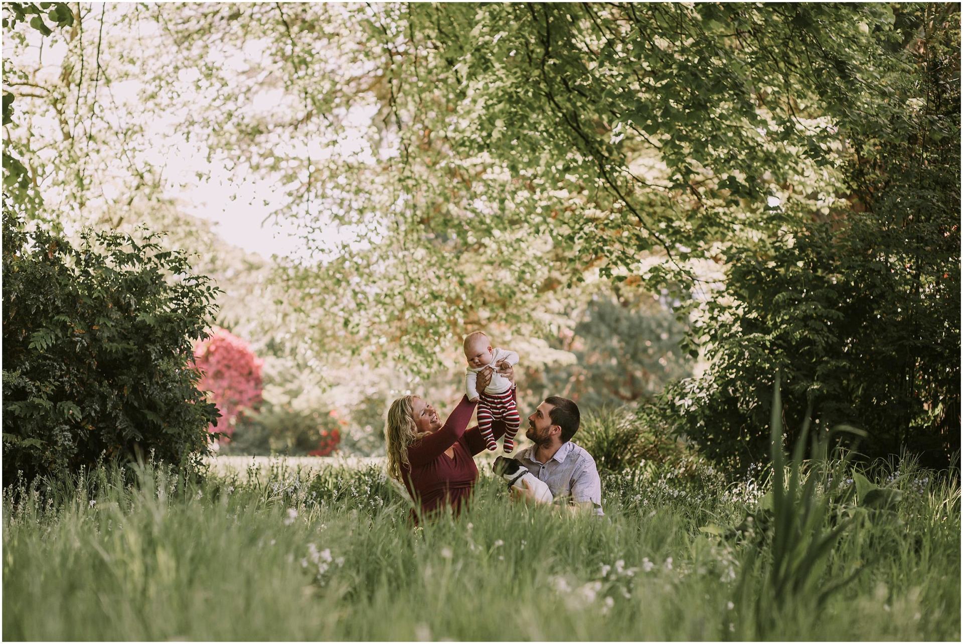 Charlotte Kiri Photography - Family Photography of a couple holding their baby up high as they look on adoringly, whilst sitting in the grass among trees and flowers in Wanaka, New Zealand.