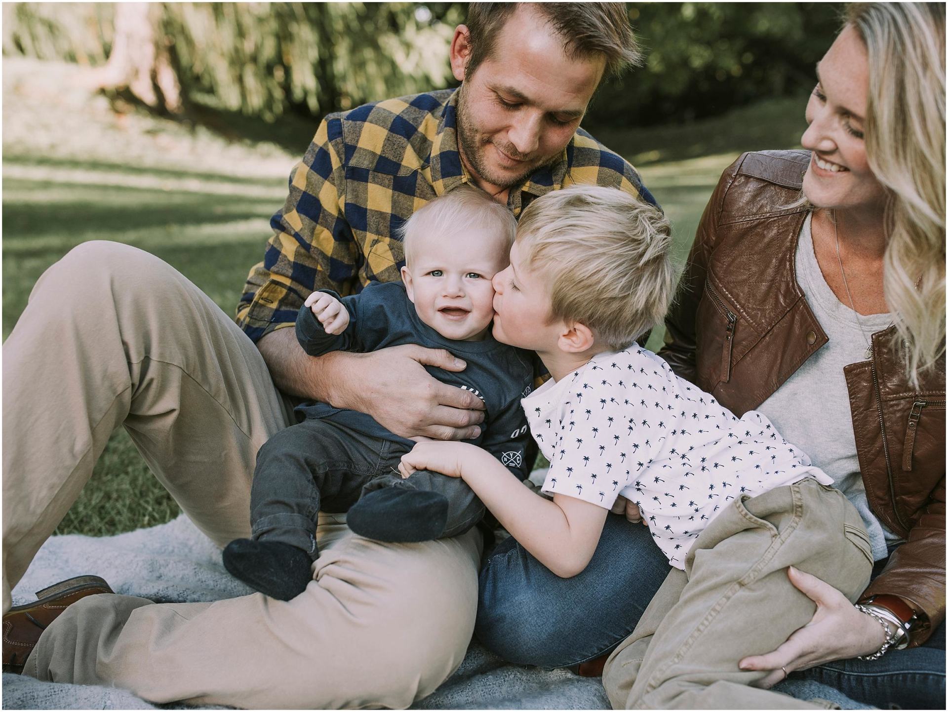 Charlotte Kiri Photography - Family Photography of a couple smiling as they watch their young son lean over to kiss his baby brother, as they sit on a picnic blanket in a garden in Wanaka, New Zealand.
