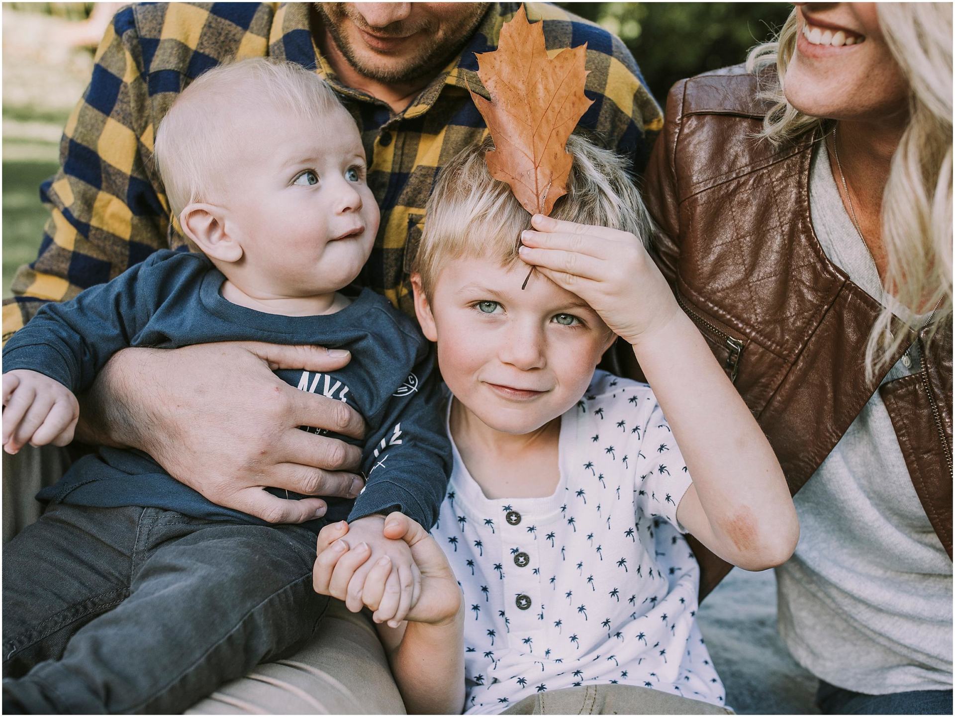 Charlotte Kiri Photography - Family Photography of a couple holding their two boys as the youngest looks up to the mother, and the eldest holds an autumn leaf up to his forehead playfully, as he holds his brother's hand in Wanaka, New Zealand.