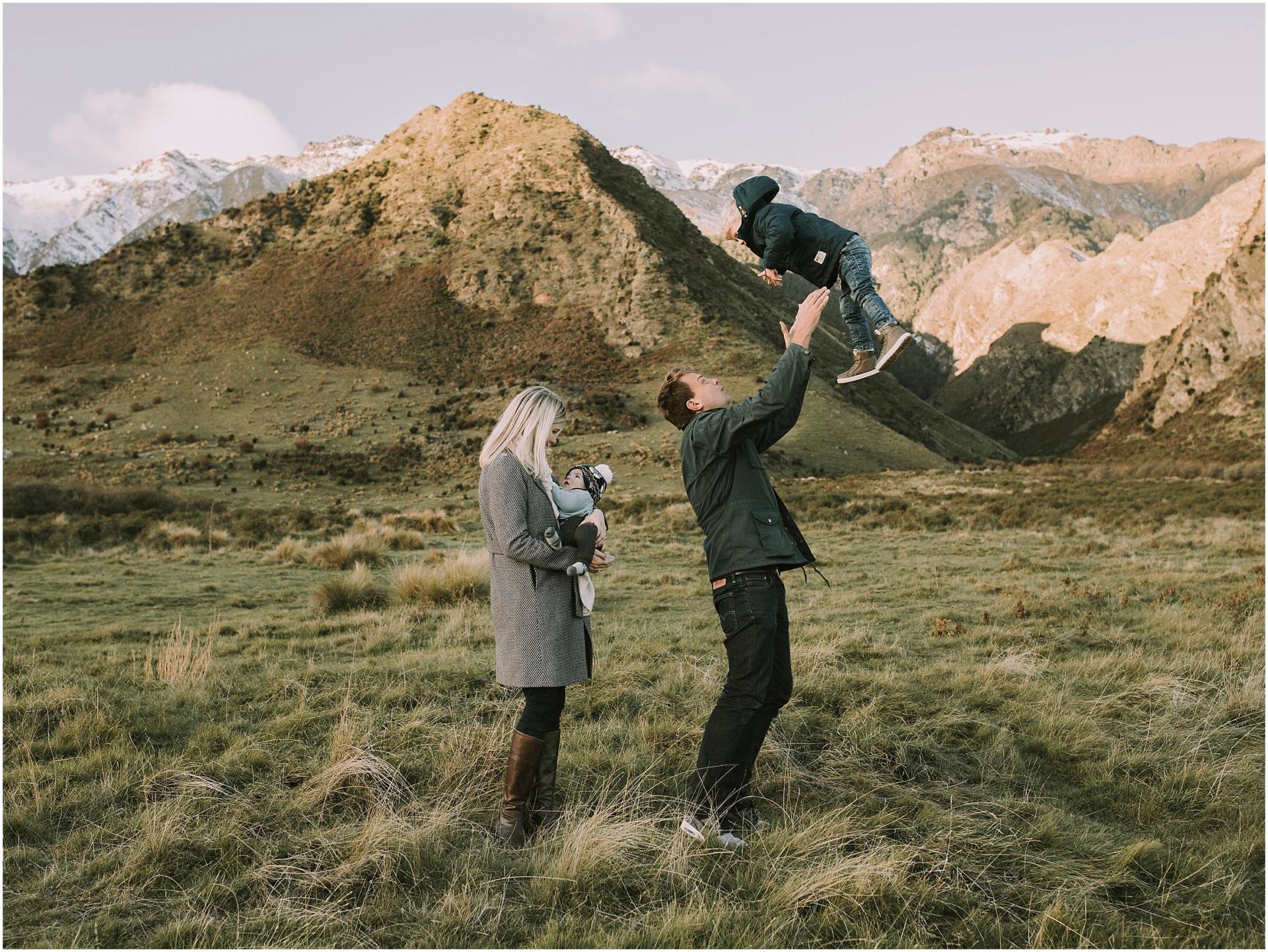 Charlotte Kiri Photography - Family Photography of a father throwing his toddler up in the air, as the mother holds her newborn baby in a field in front of mountains in Wanaka, New Zealand.