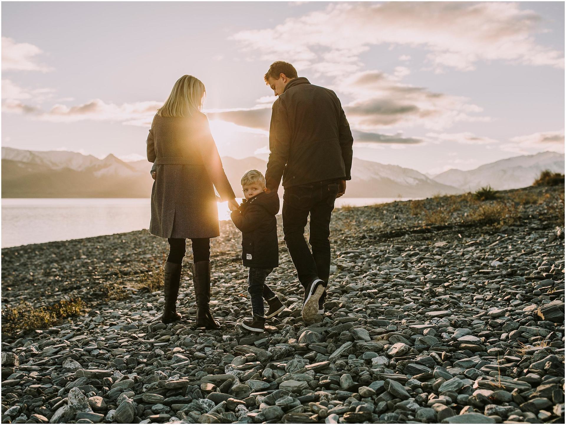 Charlotte Kiri Photography - Family Photography of a mother and father holding hands with their sweet toddler boy as he looks back, as they walk across a stony landscape in front of a lake with the sun setting behind in Wanaka, New Zealand.