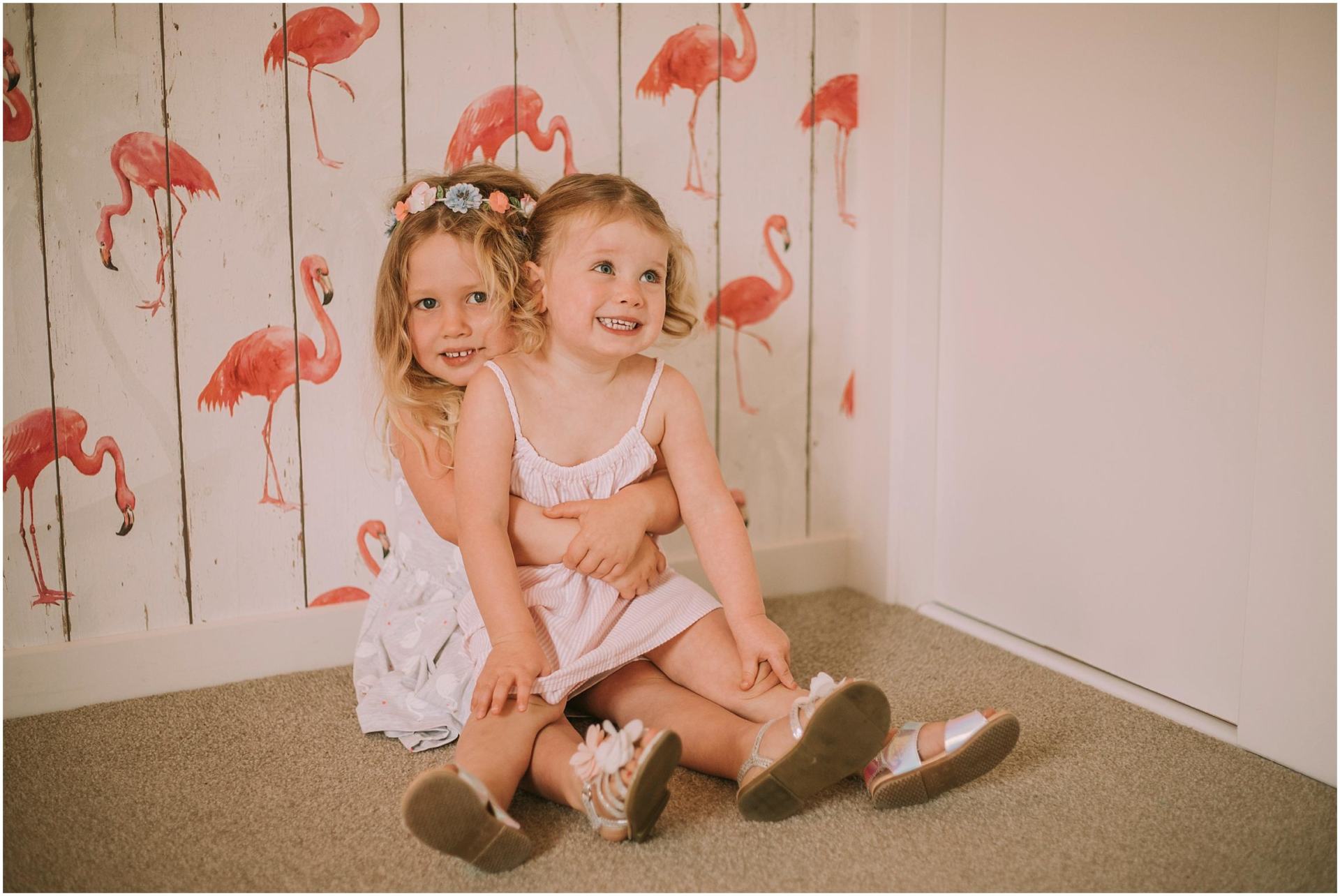 Charlotte Kiri Photography - Family Photography of a couple of young sisters hugging and smiling, as the younger girl with cute pigtails sits on her sister's lap, who wears a pretty floral wreath, in front of flamingo wallpaper in Wanaka, New Zealand.