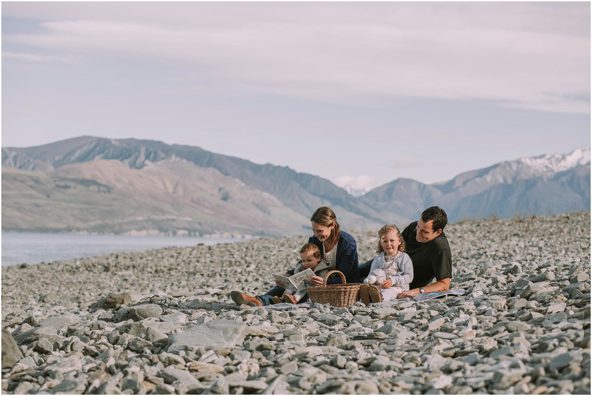 Charlotte Kiri Photography - Family Photography of a couple on a picnic blanket on the edge of a lake with mountains behind, while the mother reads to her young son, and the father looks adoringly at his daughter who is holding a soft toy in Wanaka, New Zealand.
