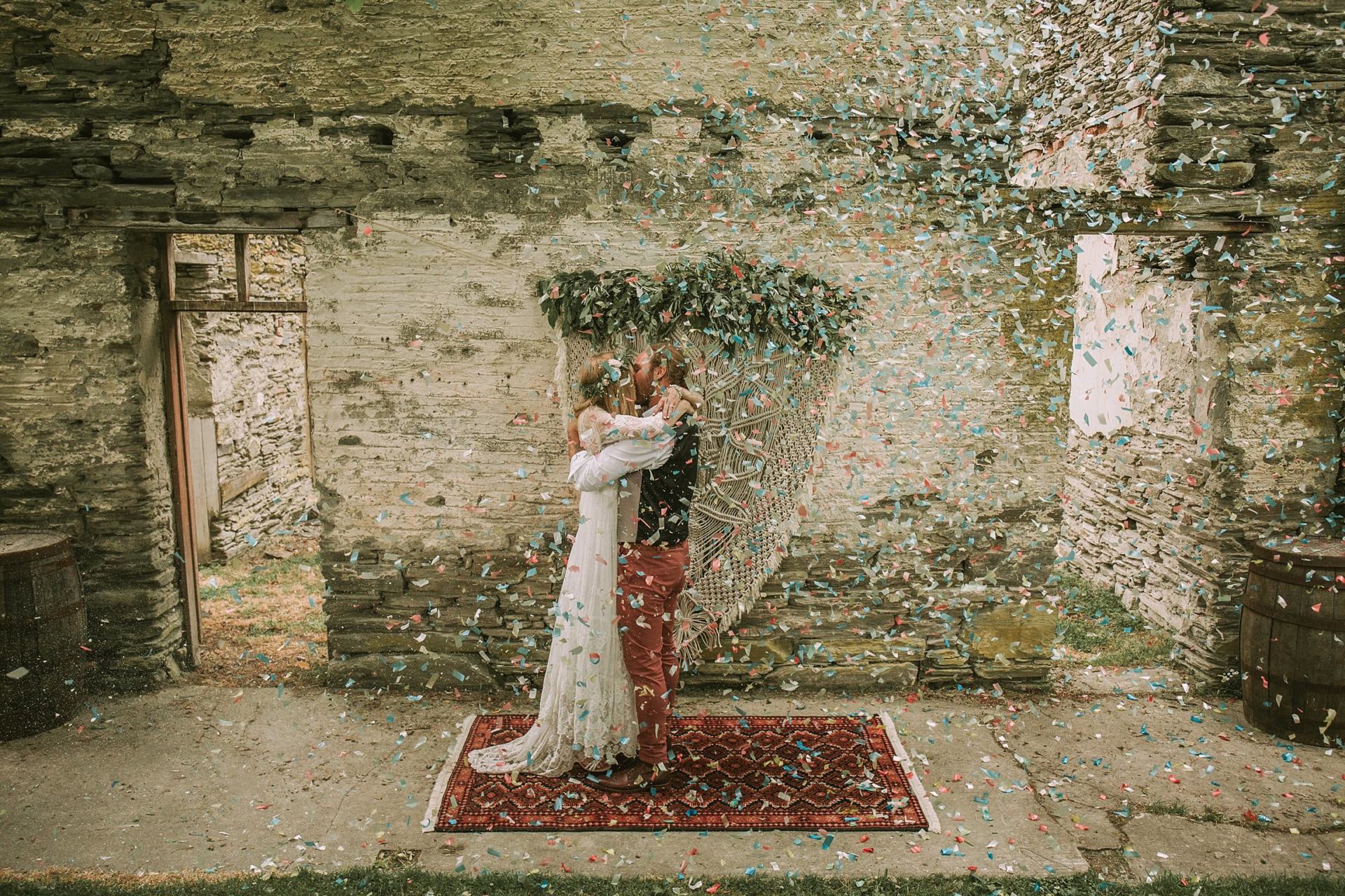 Charlotte Kiri Photography - Wedding Photography of a bohemian bride and groom, kissing and embracing as colourful confetti is thrown around them at Thurlby Domain in Arrowtown, New Zealand.