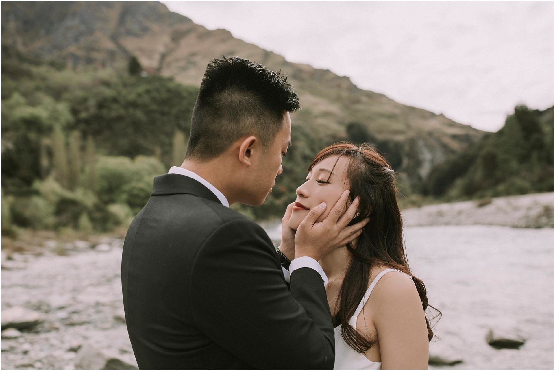Charlotte Kiri Photography - Pre-Wedding Photography with the groom wearing a smart black dinner suit looking longingly at his bride who wears a pretty shoe-string strap slip dress, as he holds her face in his hands while they stand at the water's edge with stunning landscape behind in Wanaka, New Zealand