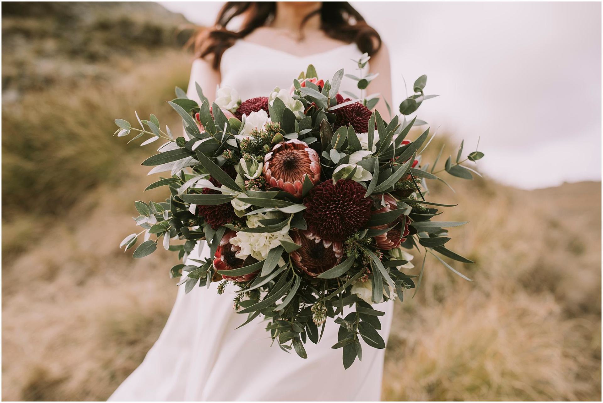 Charlotte Kiri Photography - Engagement, Pre and Post-Wedding Photography with a bride standing on a mountainside, wearing a pretty white V-necked sleeveless gown and holding a rustic bouquet with burgundy and white coloured flowers, and green foliage, in Wanaka, New Zealand
