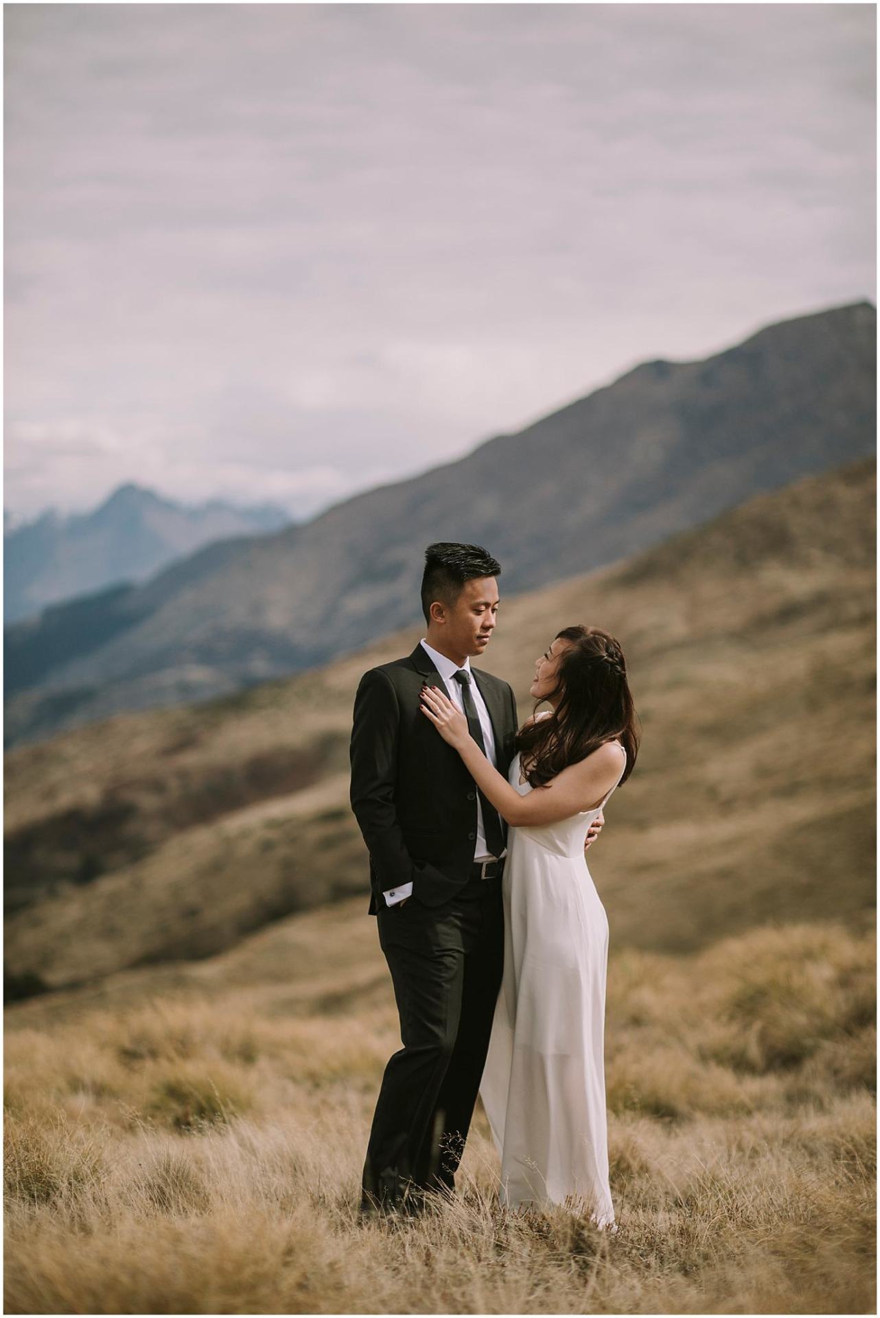 Charlotte Kiri Photography - Pre-Wedding photography with the bride wearing an elegant long cream slip gown as she puts her hand on the groom's chest who is wearing a smart black dinner suit, as they gaze at each other in a field with mountains in the background in Wanaka, New Zealand