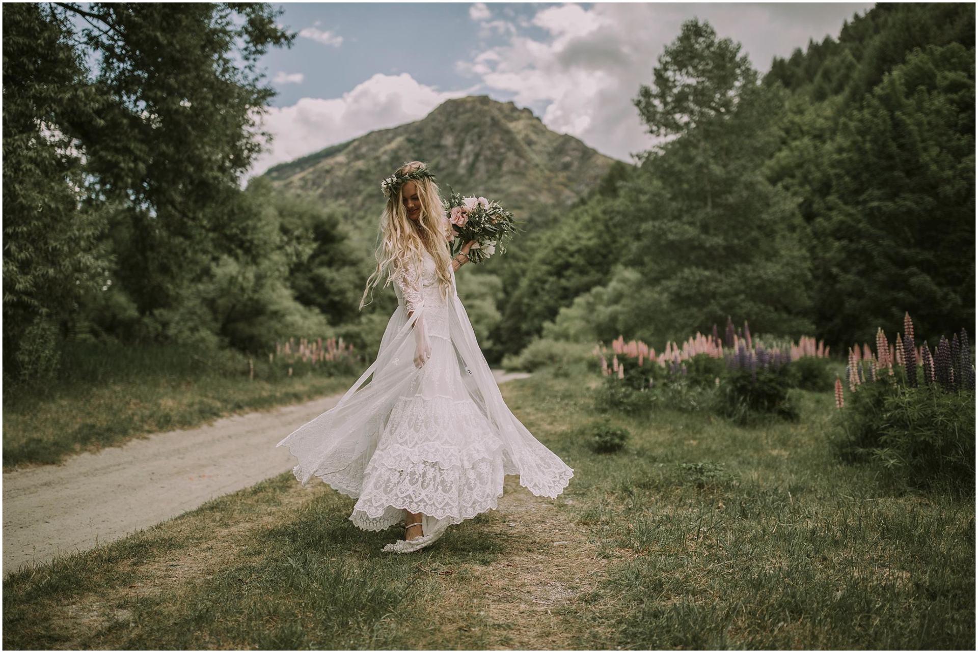 Charlotte Kiri Photography - Wedding Photography of a bride spinning around in her gorgeous bohemian lace dress, with purple and pink lupins lining a dirt road, leading to a picturesque mountain behind, in Arrowtown, New Zealand.