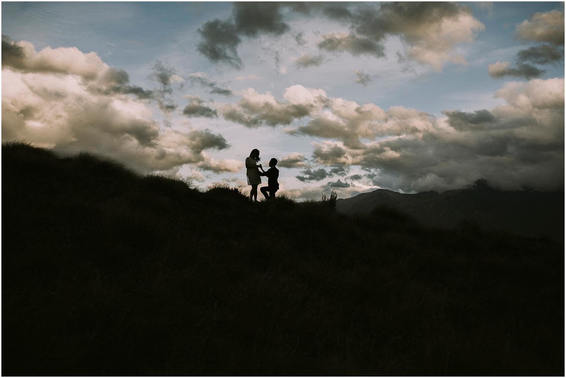 Charlotte Kiri Photography - Engagement Photography with a man kneeling proposing hopefully to his partner as they stand in silhouette on top of a mountain with moody grey and white clouds behind them in Wanaka, New Zealand
