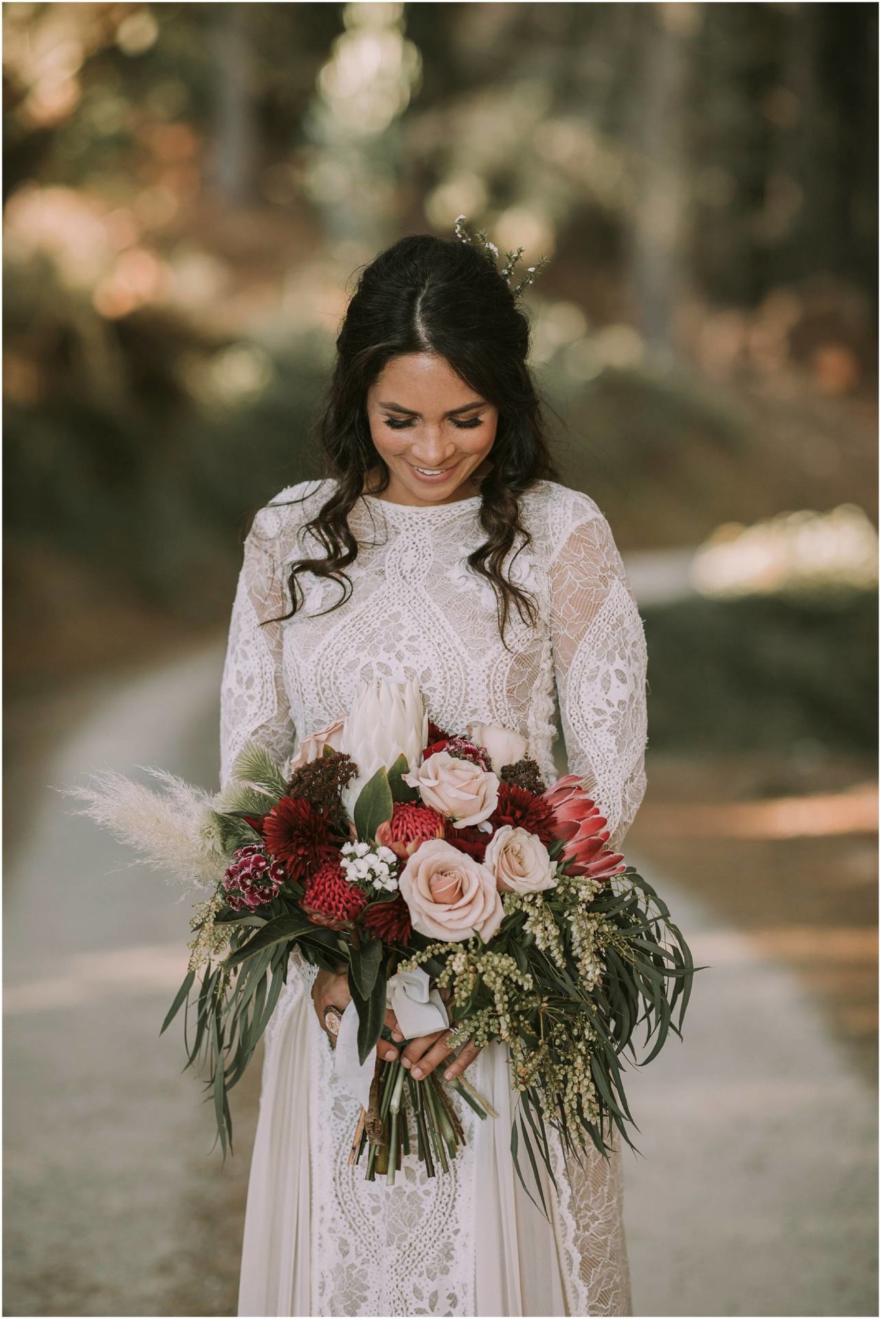Charlotte Kiri Photography - Wedding Photography of a bride wearing a stunning high-neck vintage lace gown, and looking down at her rustic bouquet as she stands on a gravel road, in Lake Hawea, New Zealand.