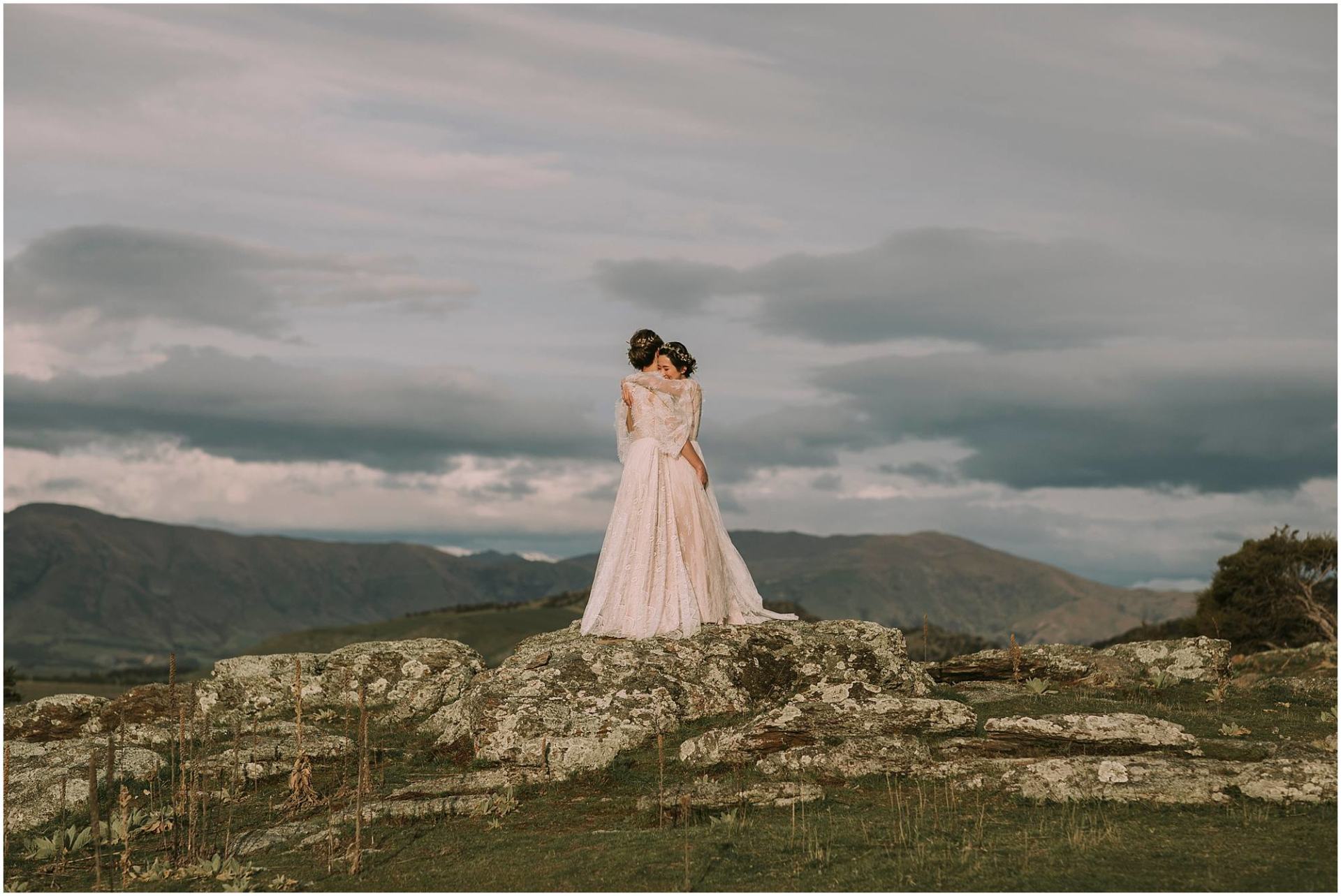 Charlotte Kiri Photography - Wedding Photography of two beautiful brides embracing on top of rocks with landscape and stormy clouds behind them
