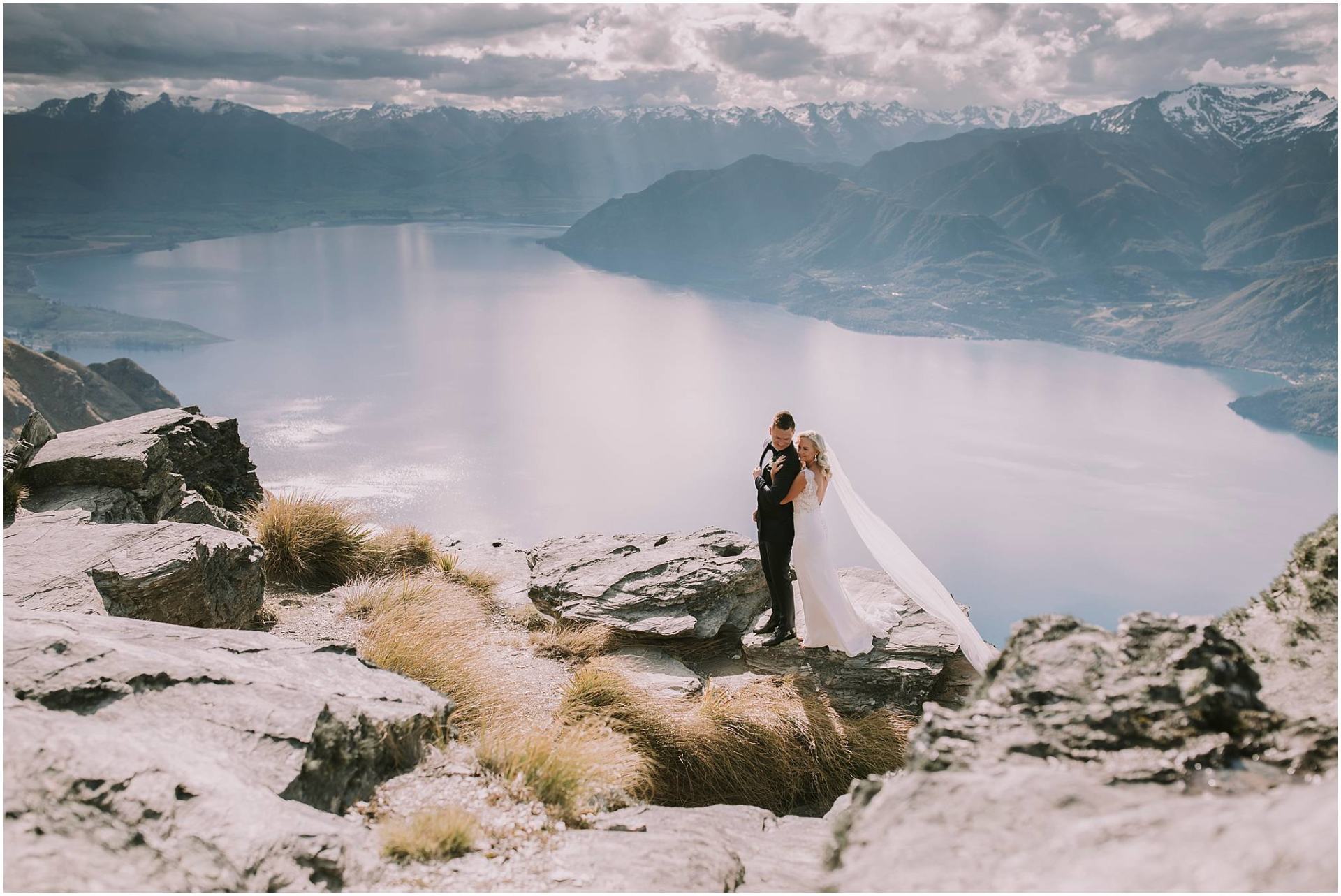 Charlotte Kiri Photography - Wedding Photography with a bride wearing a scalloped lace, low-back dress holding on to her groom, who wears a stylish black suit, as they stand on the edge of Cecil Peak with a stunning back-drop of lakes and mountains in Wanaka, New Zealand