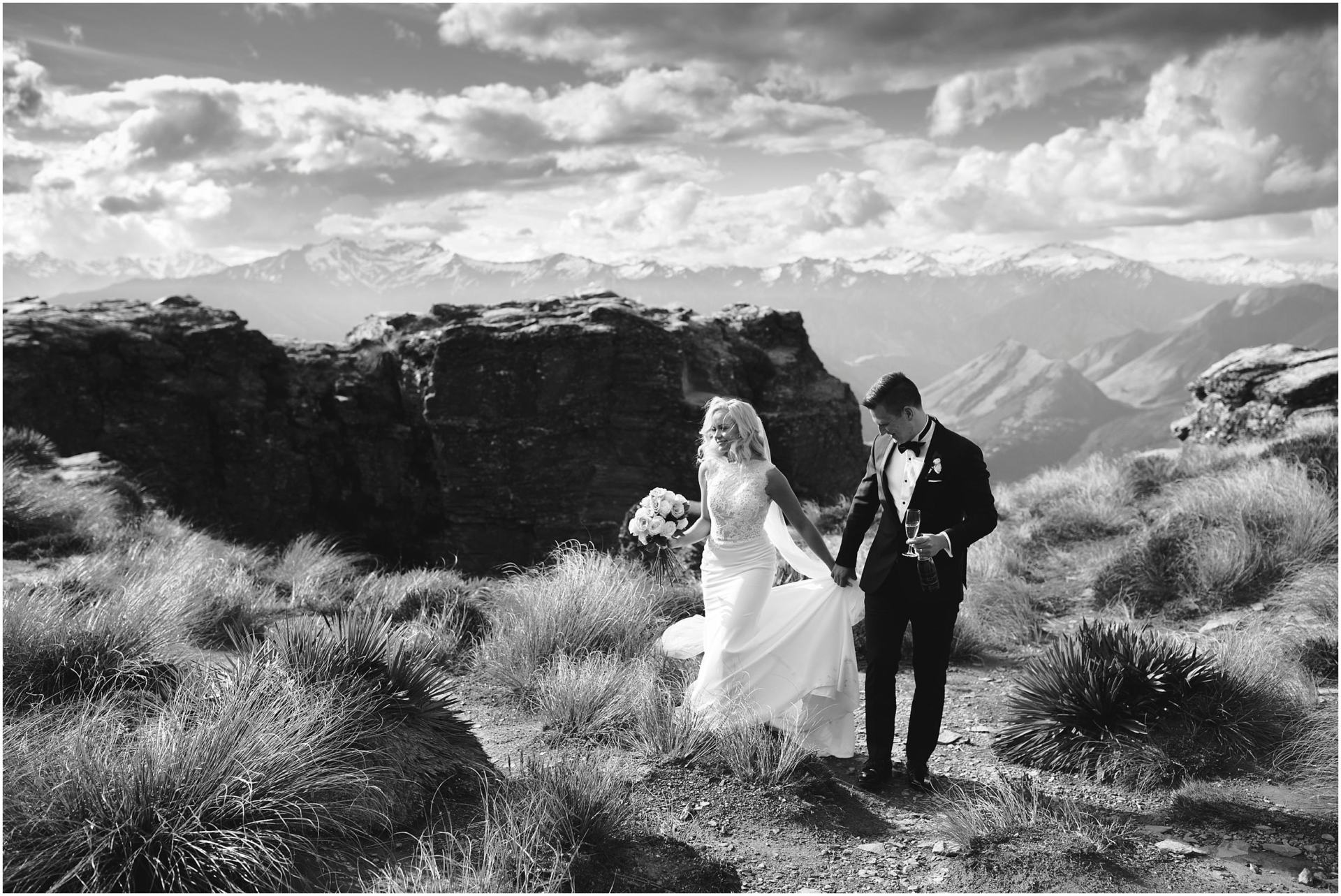 Charlotte Kiri Photography - Wedding Photography black and white picture with a bride wearing a pretty high-neck lace dress with fitted skirt, who holds her train along with her grooms hand, as they walk sipping champagne on top of Cecil Peak in Wanaka, New Zealand
