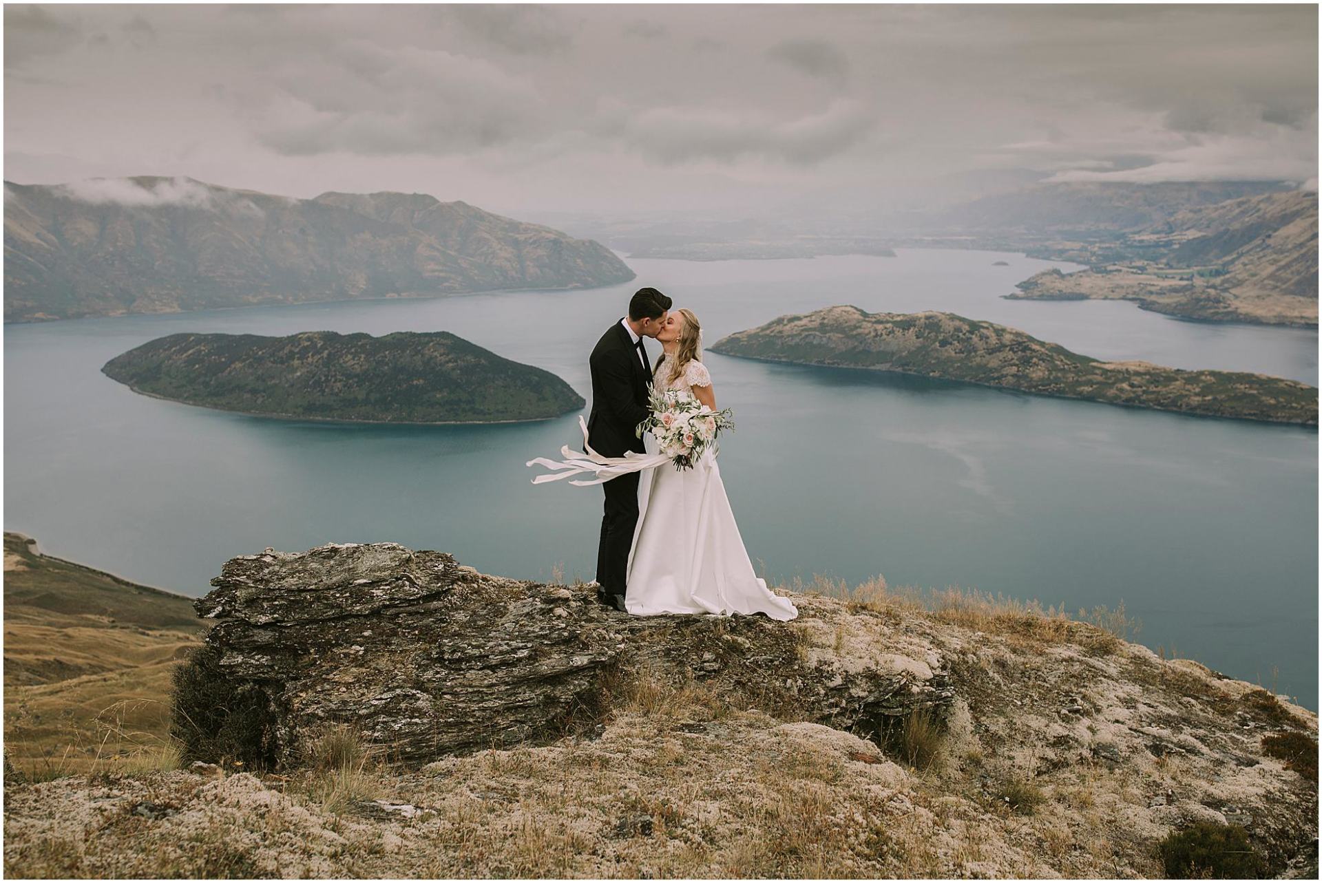 Charlotte Kiri Photography - Wedding Photography of a bride wearing a pretty lace capped-sleeved dress and holding a bouquet with ribbons that are floating in the wind, as she kisses her groom who wears a smart black suit on top of a rock that overlooks stunning lakes and mountains, in Rippon, Wanaka, New Zealand.