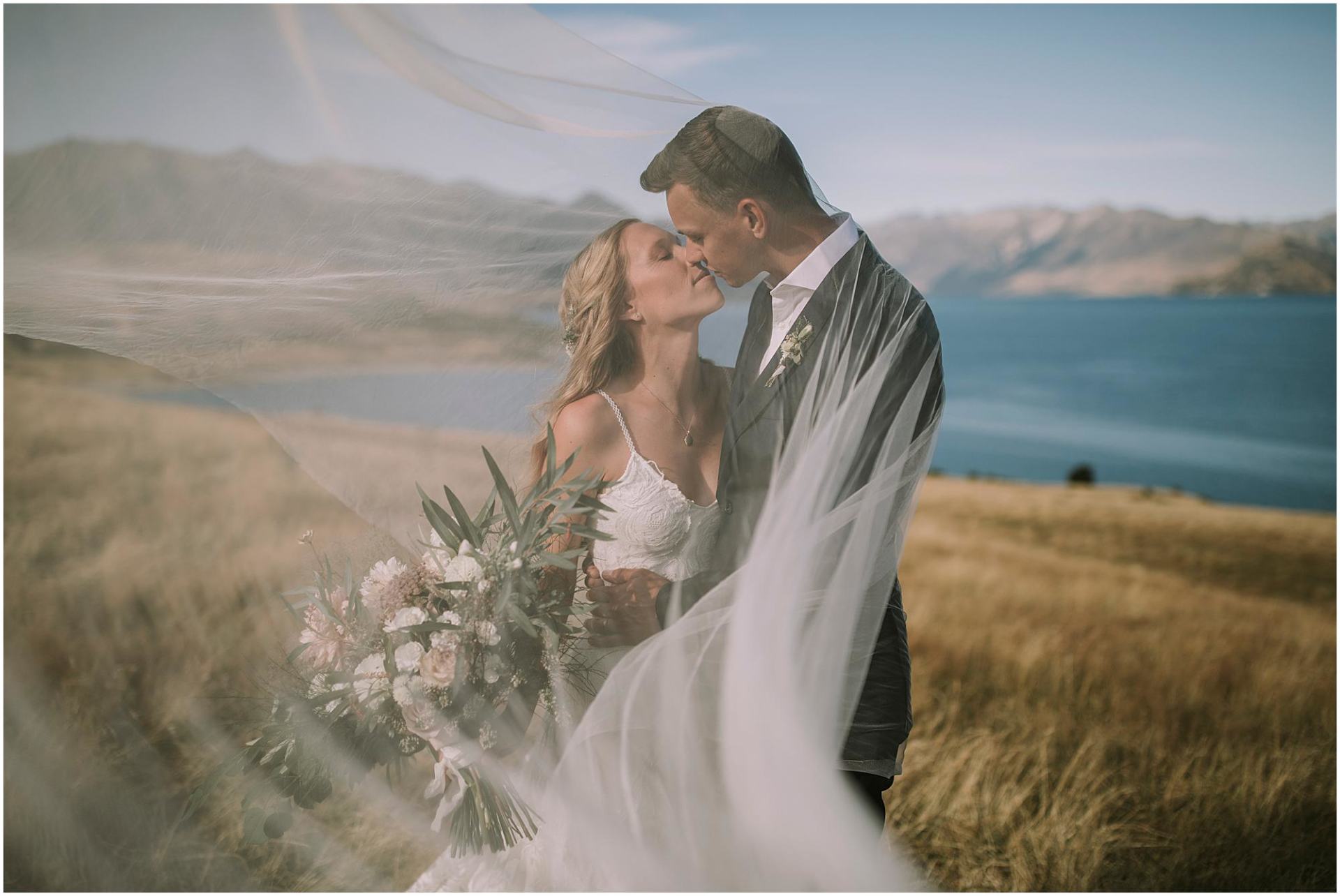 Charlotte Kiri Photography - Wedding Photography of a bride wearing a pretty shoestring strap lace dress leaning in to kiss her groom who wears a smart black suit with a white open shirt, as he holds her waist as the veil floats around them, in front of Lake Hawea in Wanaka, New Zealand