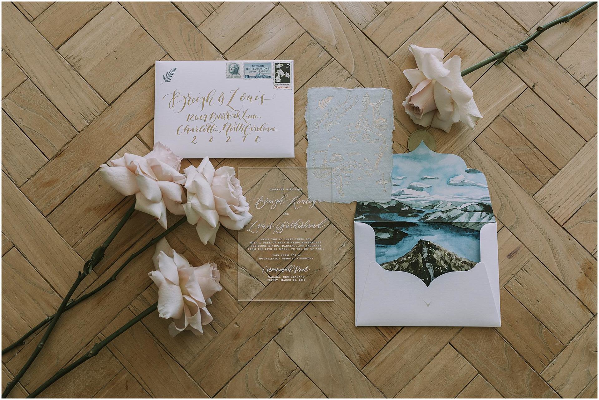Charlotte Kiri Photography - Wedding Photography of pink roses scattered across stylish wedding invitations with fern emblems, and gold and white calligraphy, accompanied by lovely envelopes with Wanaka scenery of mountains and lakes printed inside them, in Wanaka, New Zealand.