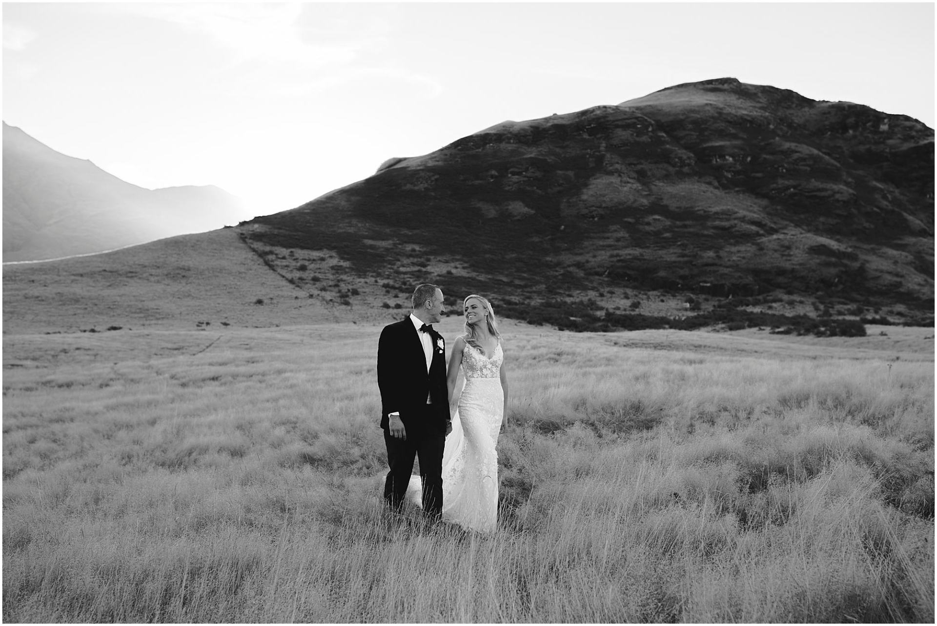 Charlotte Kiri Photography - Wedding Photography black and white photo of a bride wearing a lovely floral lace fitted dress, and her groom wearing a classy black suit, walking hand in hand as they cross over a grassy plain, with mountains behind in Wanaka, New Zealand.