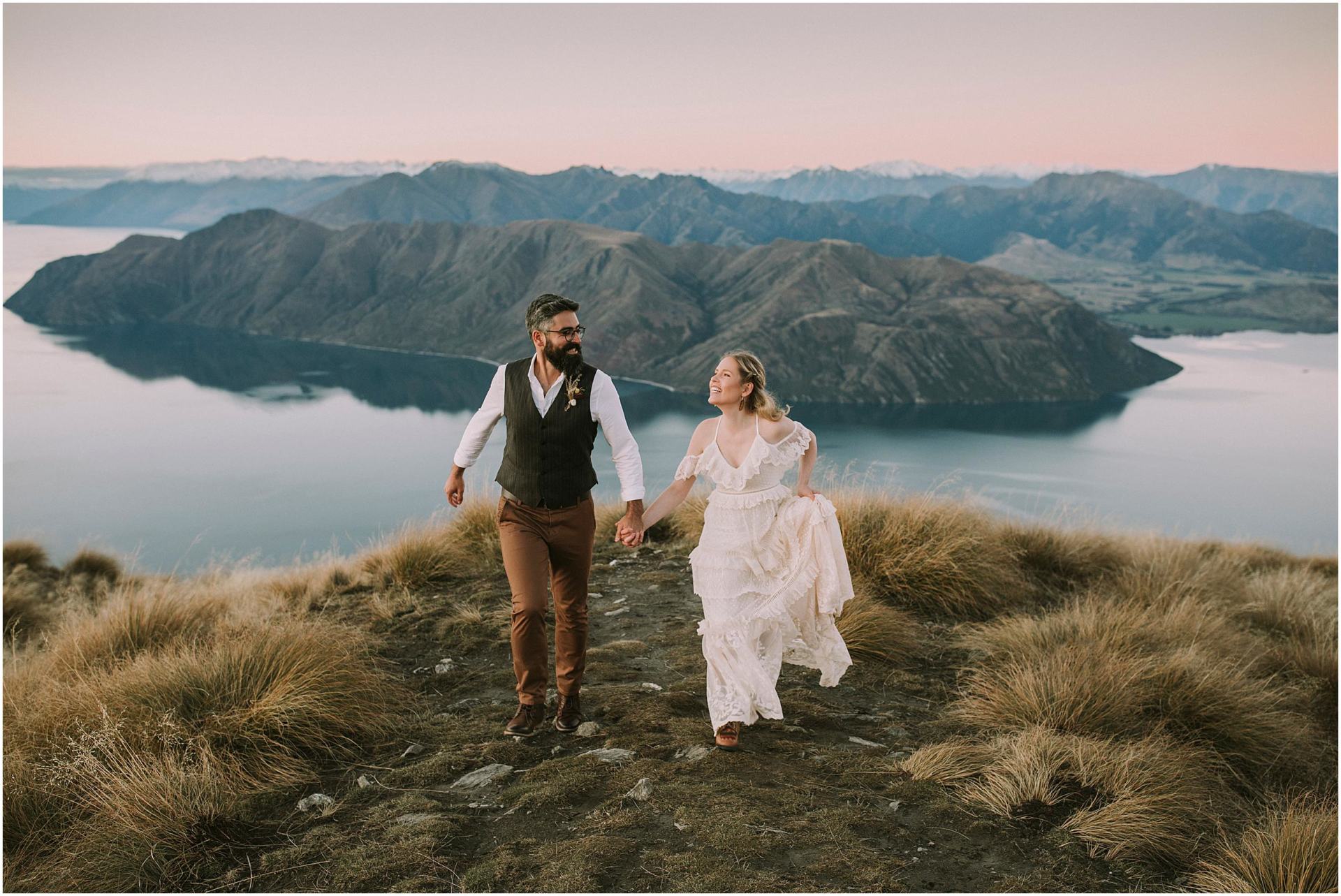 Charlotte Kiri Photography - elopement photography with the bride and groom holding hands and laughing, on top of Roy's Peak, with a stunning view of lakes and mountains behind them in Wanaka, New Zealand