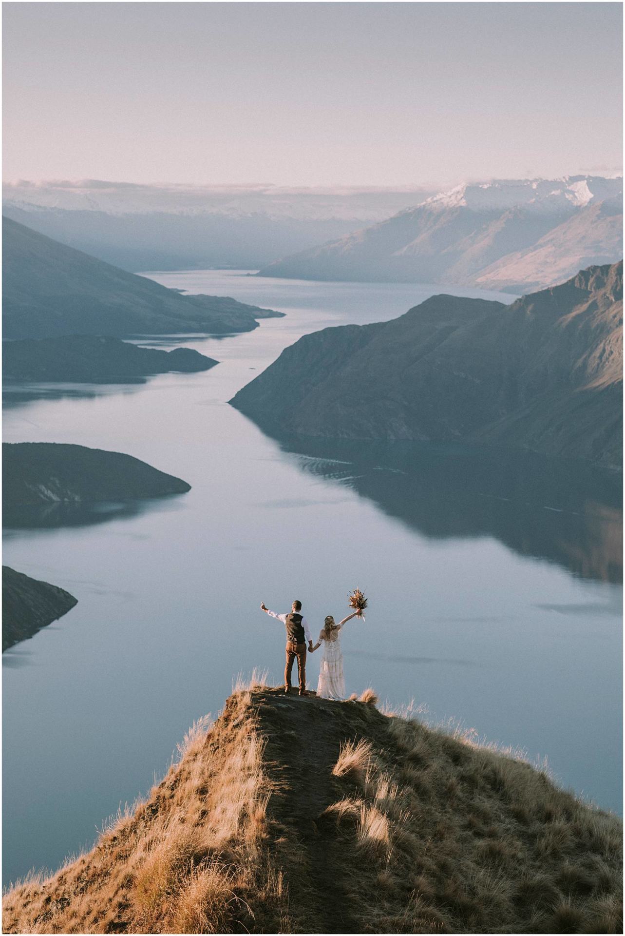 Charlotte Kiri Photography - elopement photography with bride and groom standing on top of Roy's Peak with arms outstretched with glacial lakes in front, Wanaka New Zealand