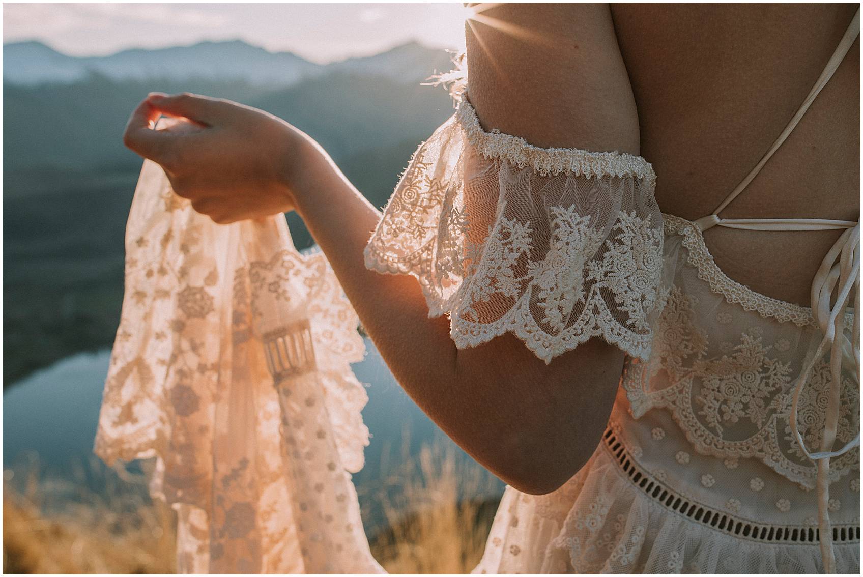 Charlotte Kiri Photography - elopement photography showing the back of a bride's dress with vintage lace and off-shoulder details, the bride is holding her train with the view of lakes, mountains and sunsets shining through in the background