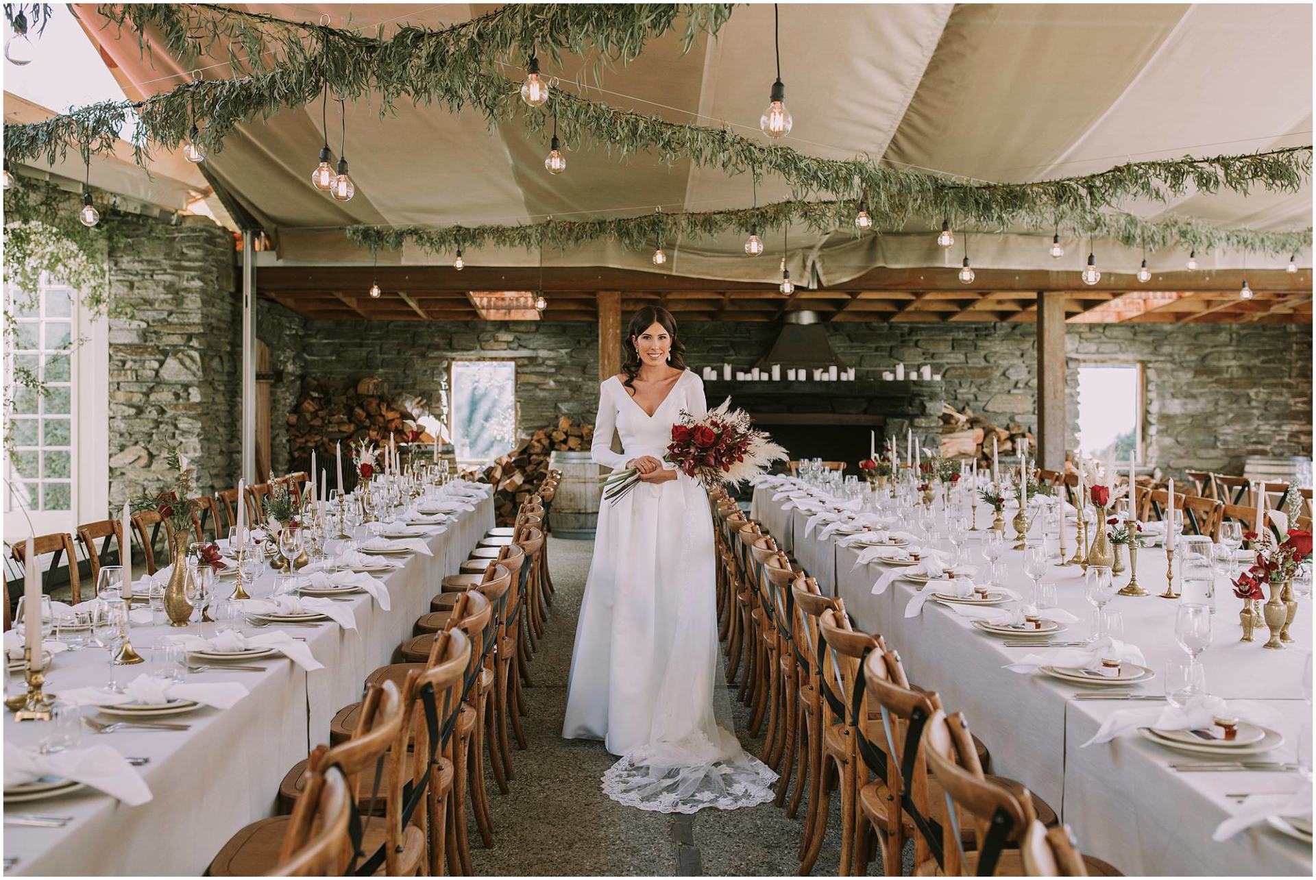 Charlotte Kiri - Wedding Photography of a bride wearing an elegant long-sleeved V-necked dress with a sheer floral lace veil, and holding a dark mahogany floral and toi toi bouquet amidst long tables, wooden chairs, and formal settings at the reception in Queenstown, New Zealand.