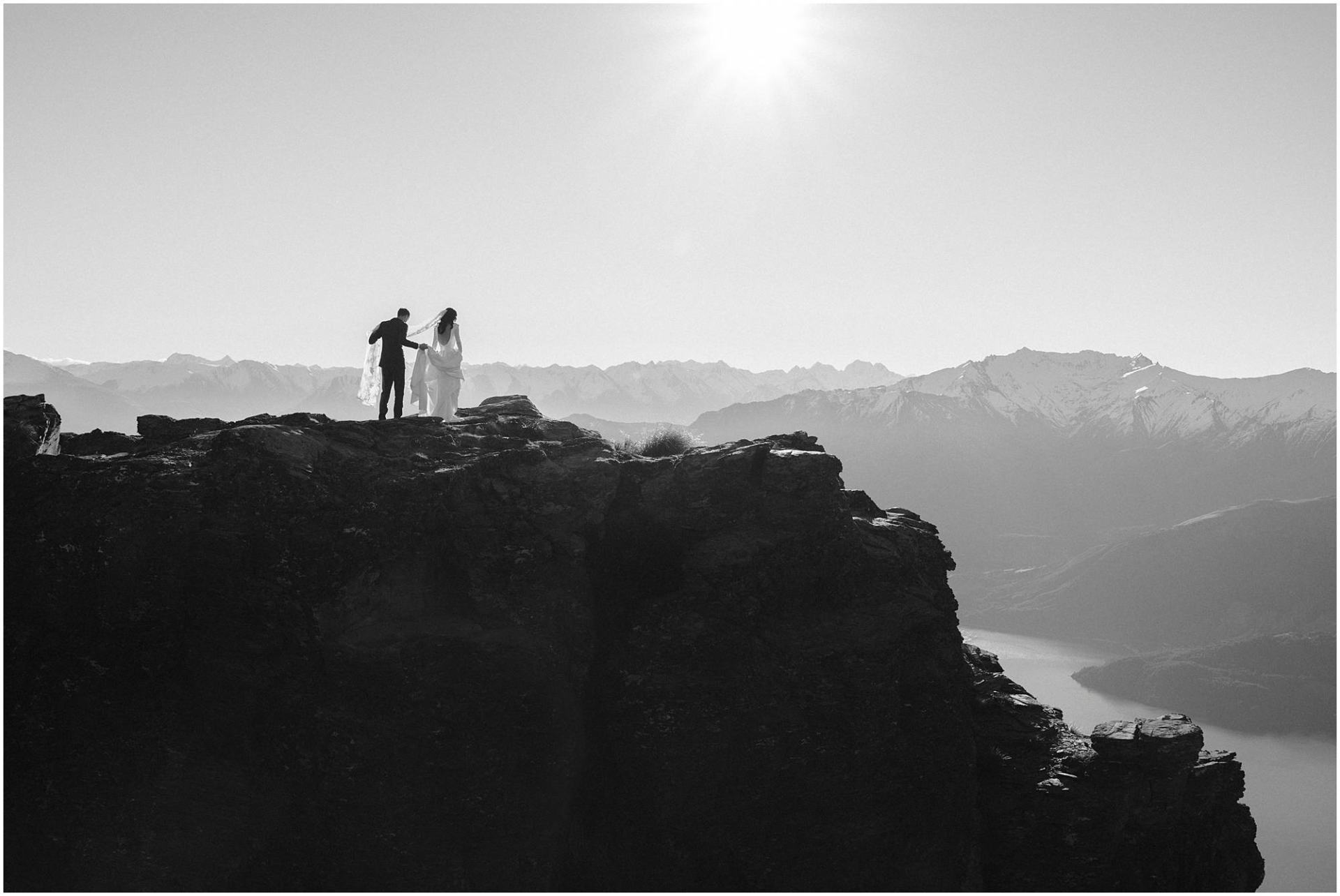 Charlotte Kiri Photography - Wedding Photography black and white photo of an elegant bride, and groom who holds her train and veil, as they walk towards the end of a cliff, with a spectacular view of glacial lakes and mountains in front of them, in Wanaka, New Zealand.