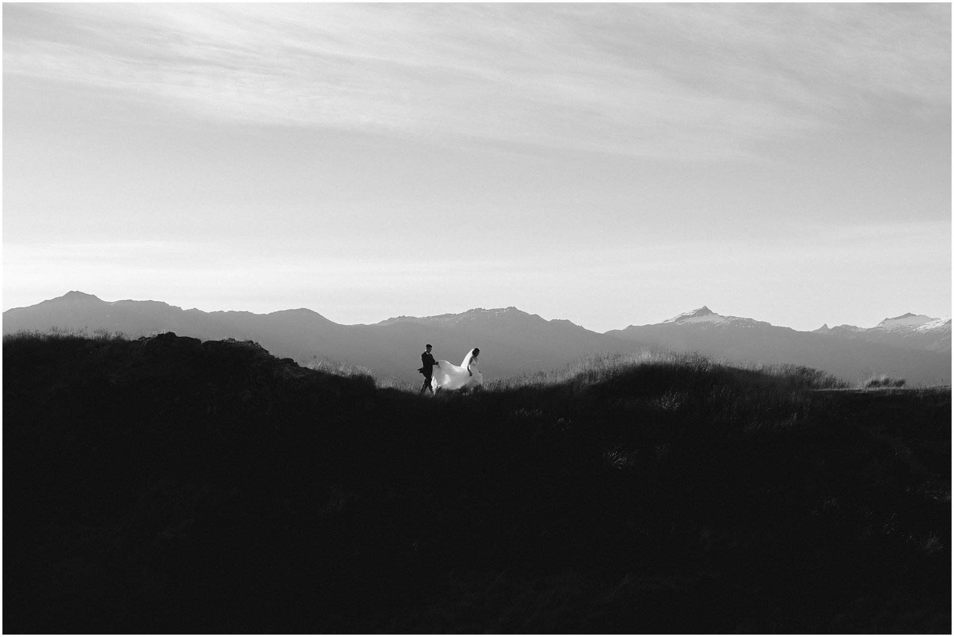 Charlotte Kiri Photography - Pre and Post Wedding Photography with a black and white image of an elegant bride walking in front of her groom across a peak, as he attentively holds her long train over the grass, with a silhouette of mountainous terrain behind them, in Wanaka, New Zealand