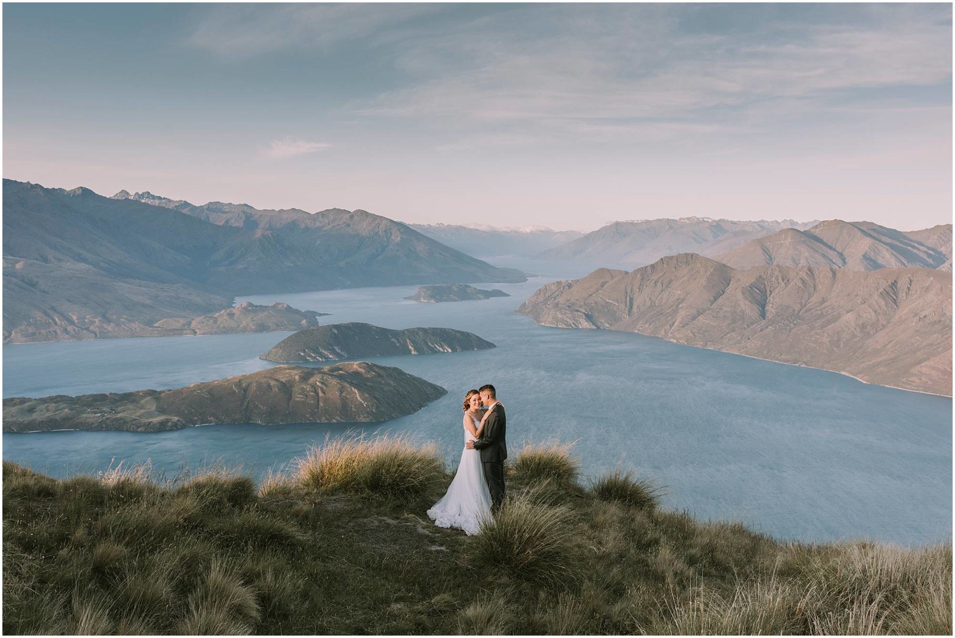 Charlotte Kiri Photography - Pre & Post Wedding Photography breathtaking image of a smiling couple embracing on the edge of Roys Peak, with stunning scenery of lakes and mountains behind them, in Wanaka, New Zealand
