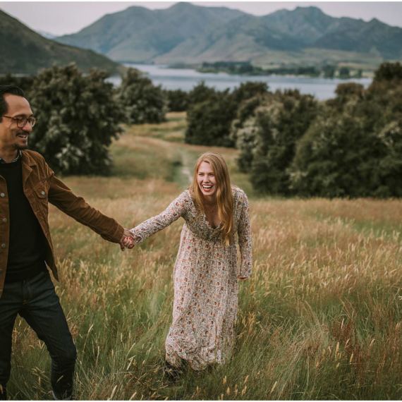 Charlotte Kiri Photography - Engagement Photography with the happy couple holding hands and smiling in a field in front of beautiful lakes and mountains in Wanaka, New Zealand
