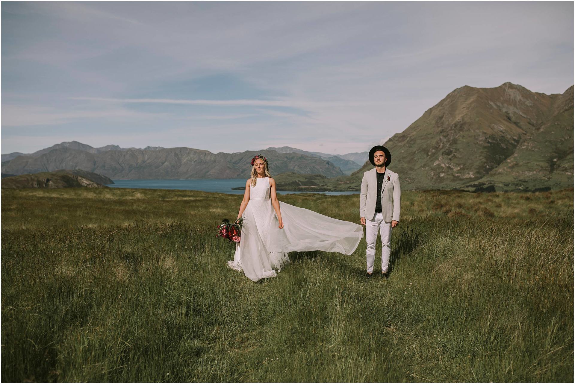 Charlotte Kiri Photography - Wedding Photography with a bride wearing a classic high-necked sleeveless gown with a flowing train, standing in a field with her husband who wears a beige suit jacket, white pants, and a black shirt and hat as they close their eyes and soak in the special moment together, with a gorgeous backdrop of mountains and lakes in Wanaka, New Zealand