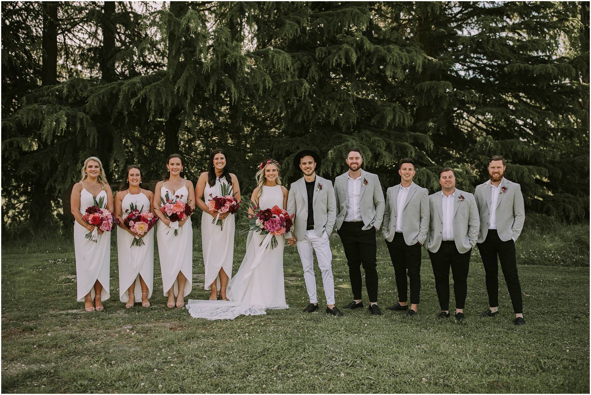 Charlotte Kiri Photography - Wedding Photography of a gorgeous bridal party, with the bride and bridesmaids wearing crisp white dresses with red, pink, and peach bouquets, and the groom wearing smart white pants, a grey jacket, and black shirt and hat, alongside his groomsmen with black pants, white shirts, and grey jackets, as they pose in front of a line of trees. in Wanaka, New Zealand